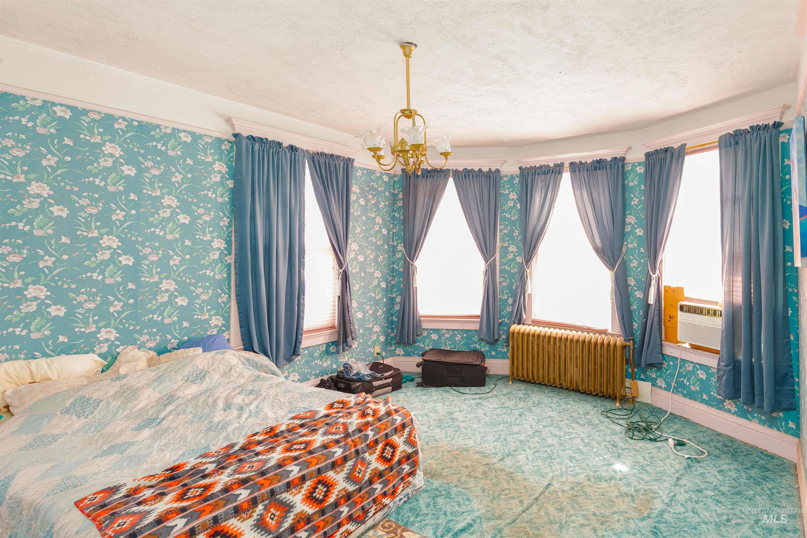 Carpeted bedroom with a chandelier, radiator heating unit, a textured ceiling, and wallpapered walls