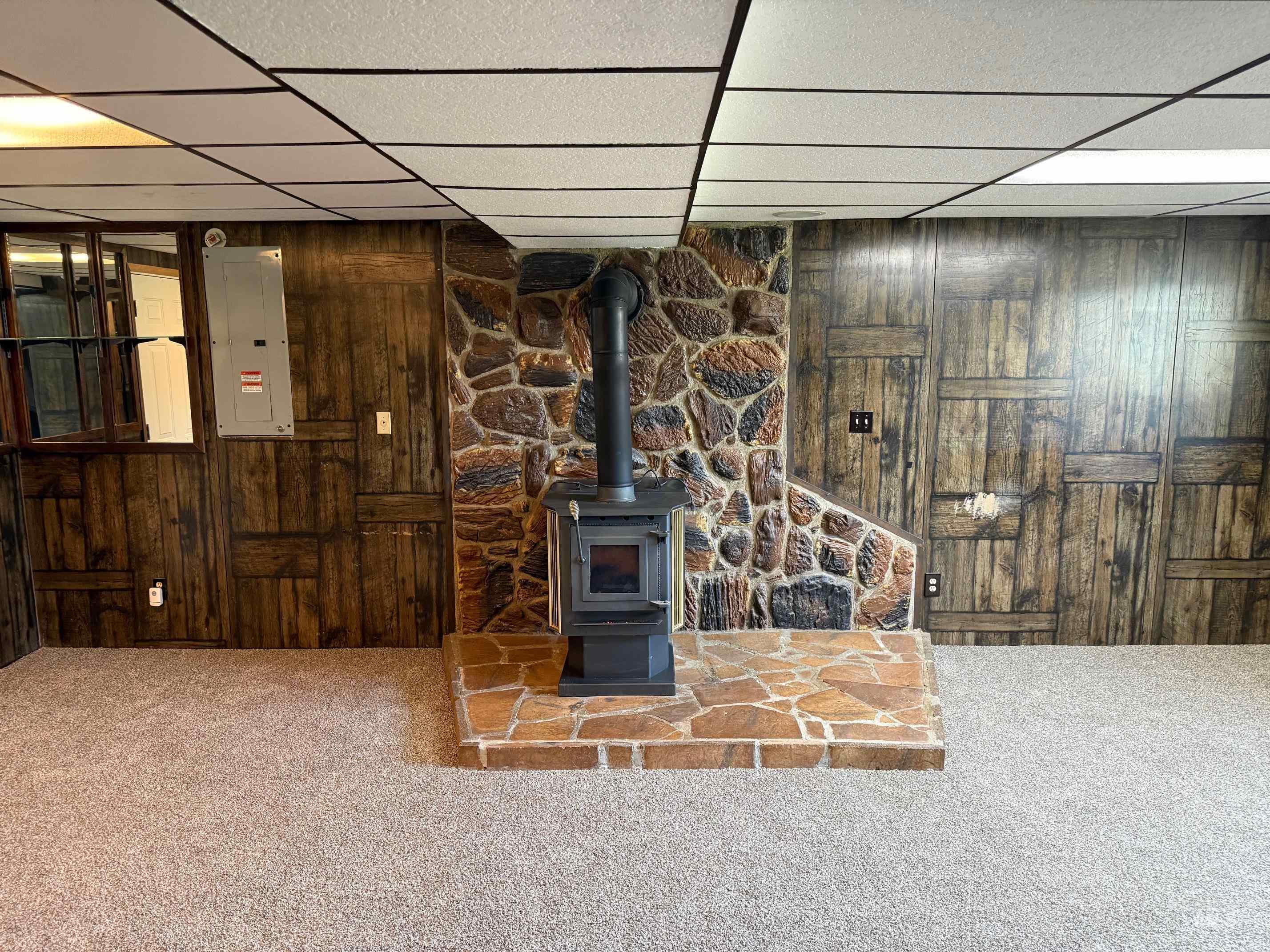Unfurnished living room featuring wooden walls, a wood stove, carpet flooring, a drop ceiling, and electric panel