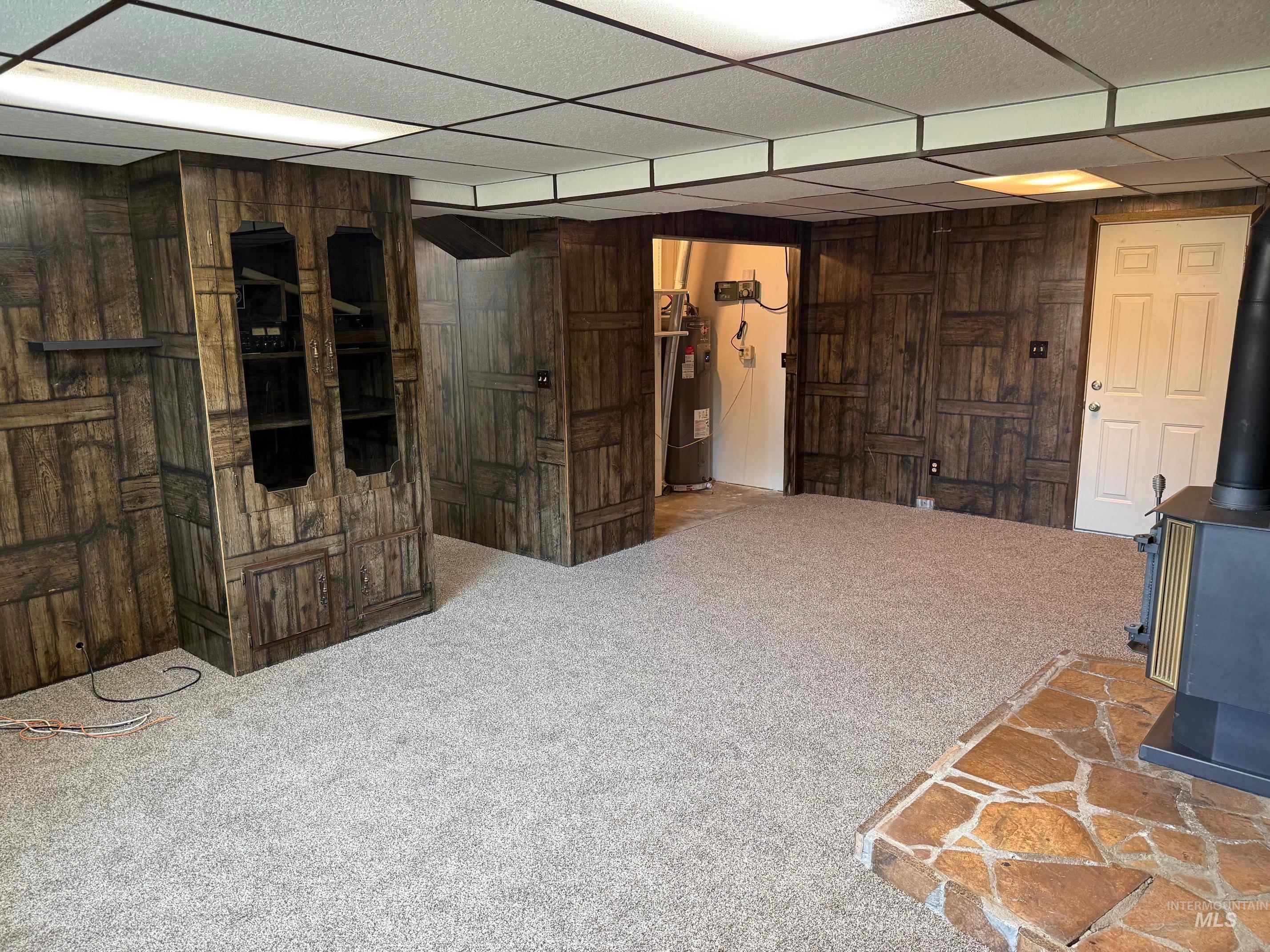 Finished basement featuring a paneled ceiling, carpet floors, wooden walls, a wood stove, and water heater