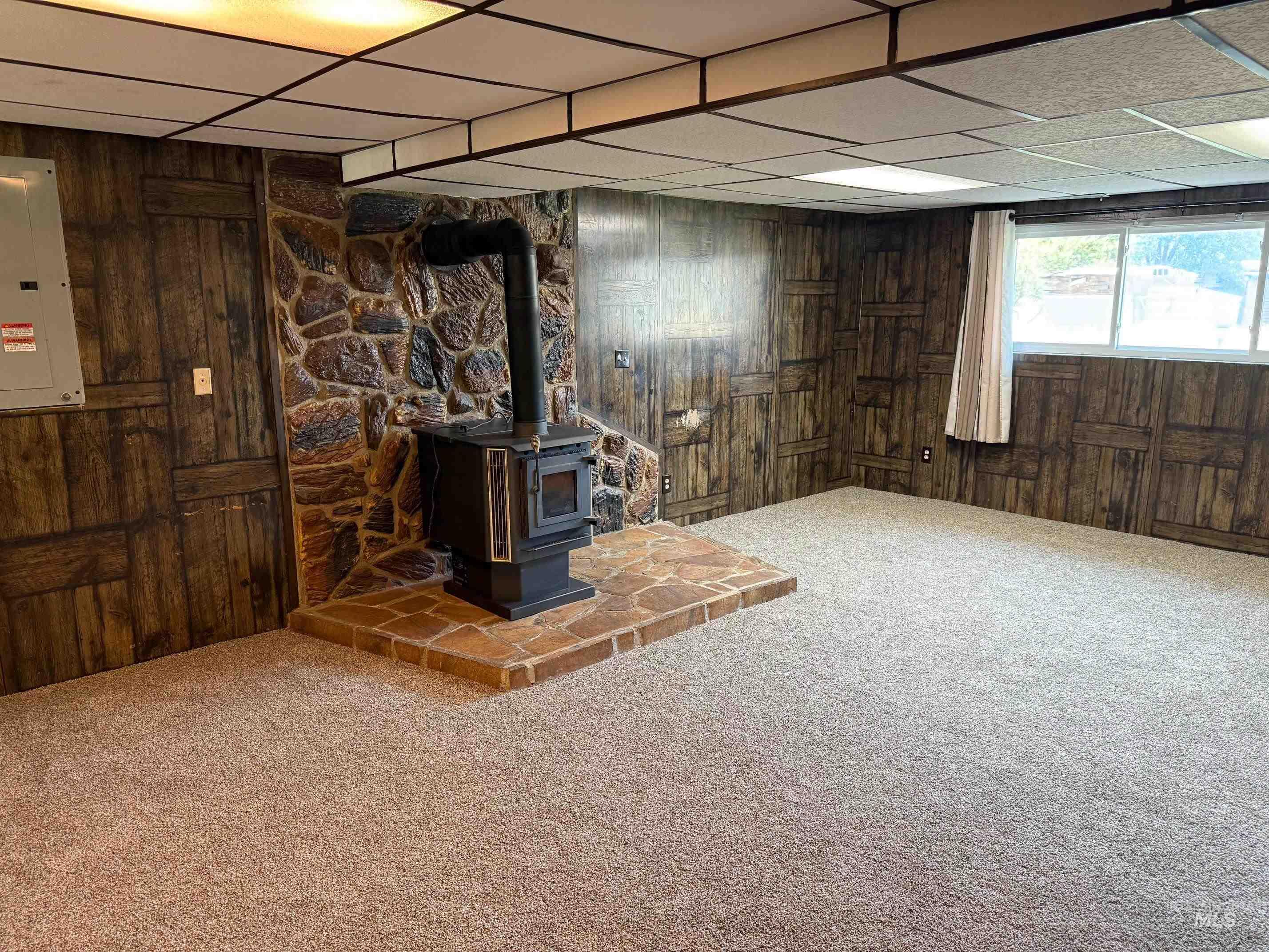 Below grade area featuring wood walls, a wood stove, carpet floors, a drop ceiling, and electric panel