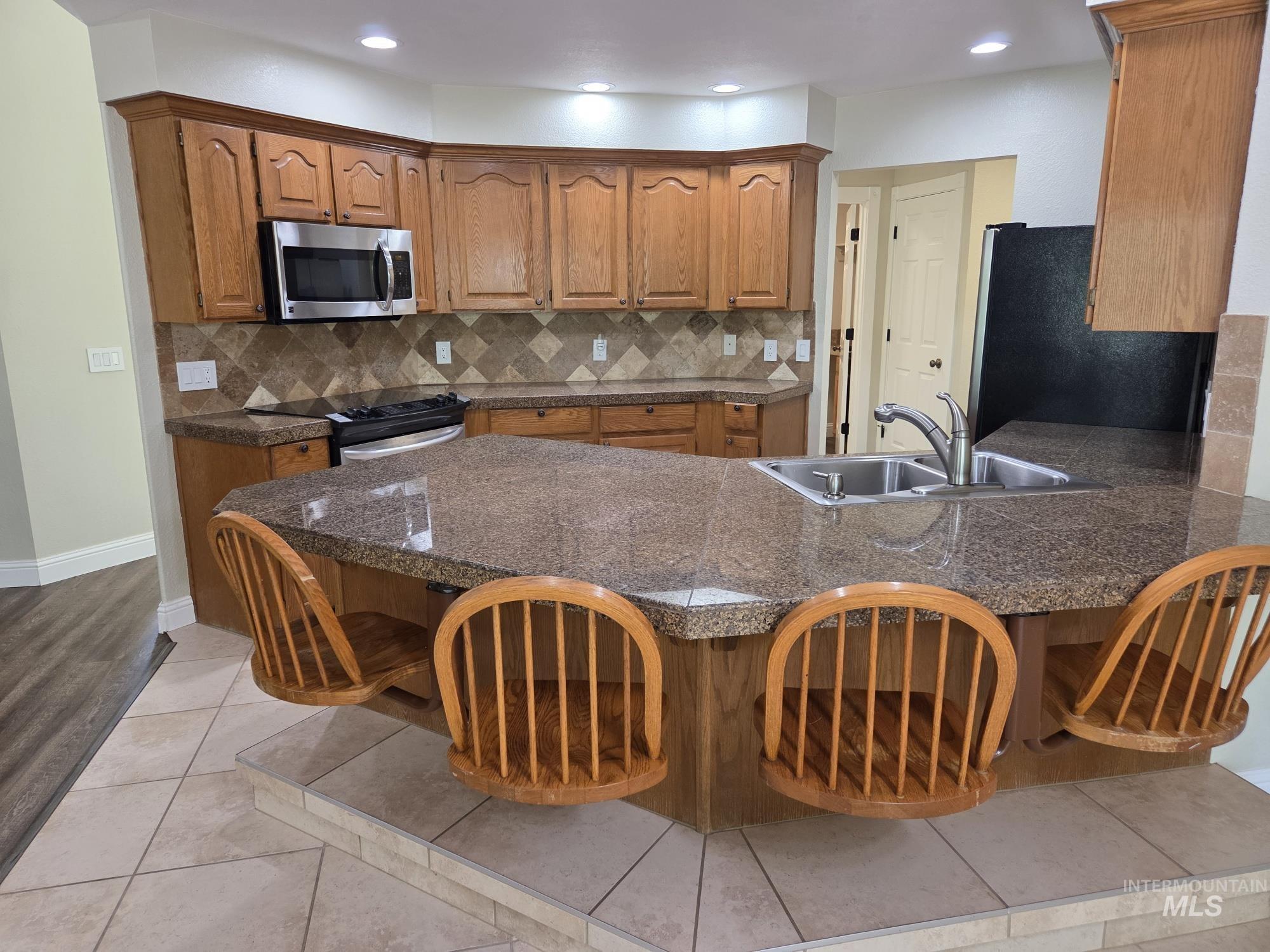 Kitchen featuring tile countertops, backsplash, a peninsula, brown cabinetry, and recessed lighting