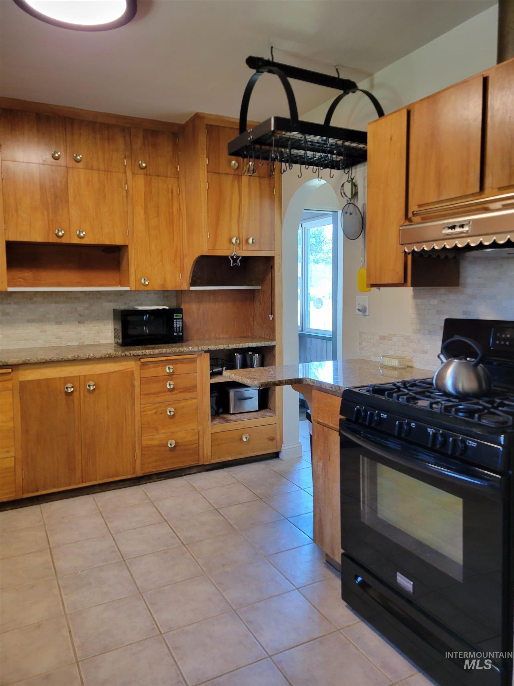 Kitchen featuring backsplash, black appliances, light tile patterned floors, range hood, and brown cabinets