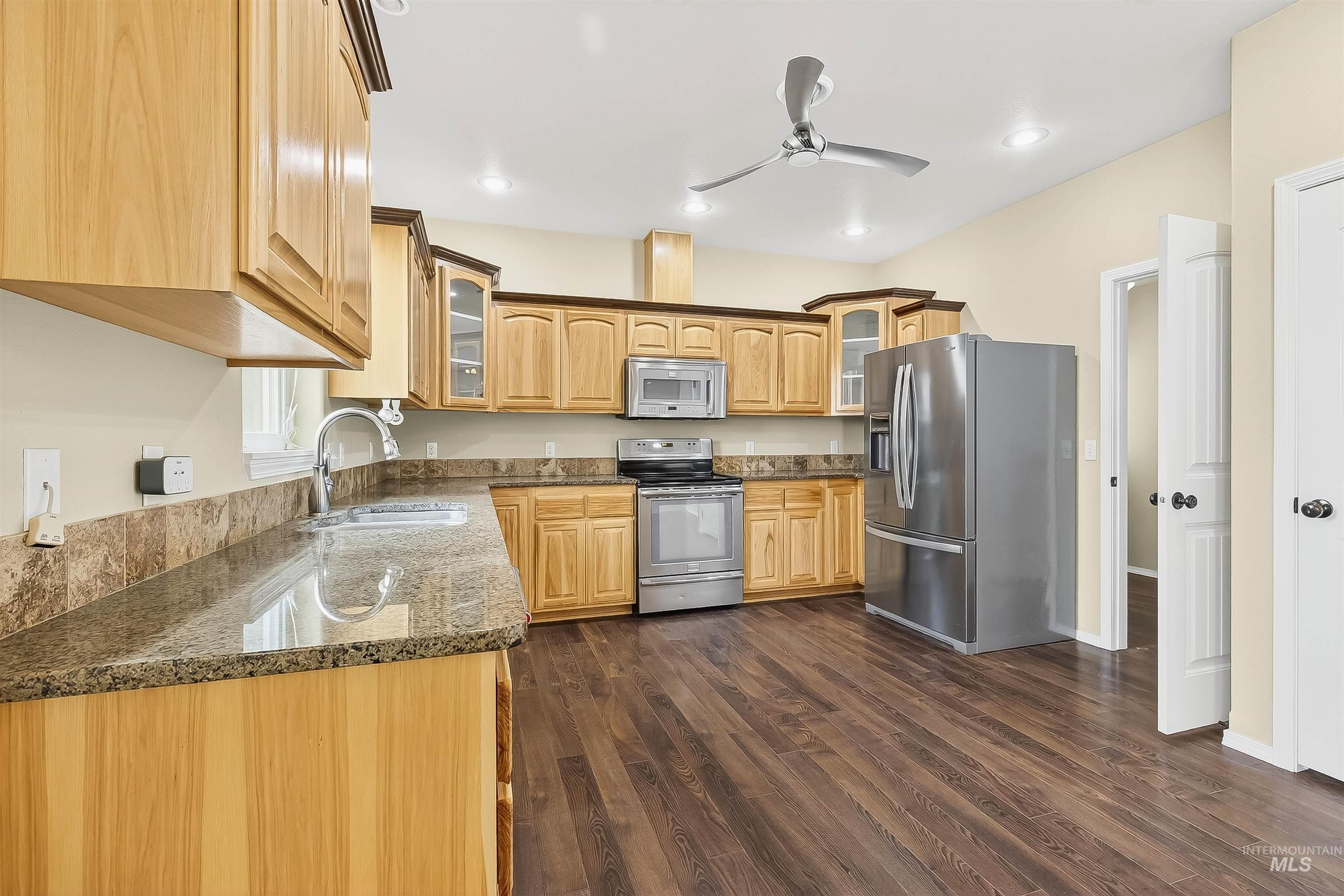 Kitchen featuring glass insert cabinets, appliances with stainless steel finishes, dark stone counters, dark wood-style floors, and ceiling fan
