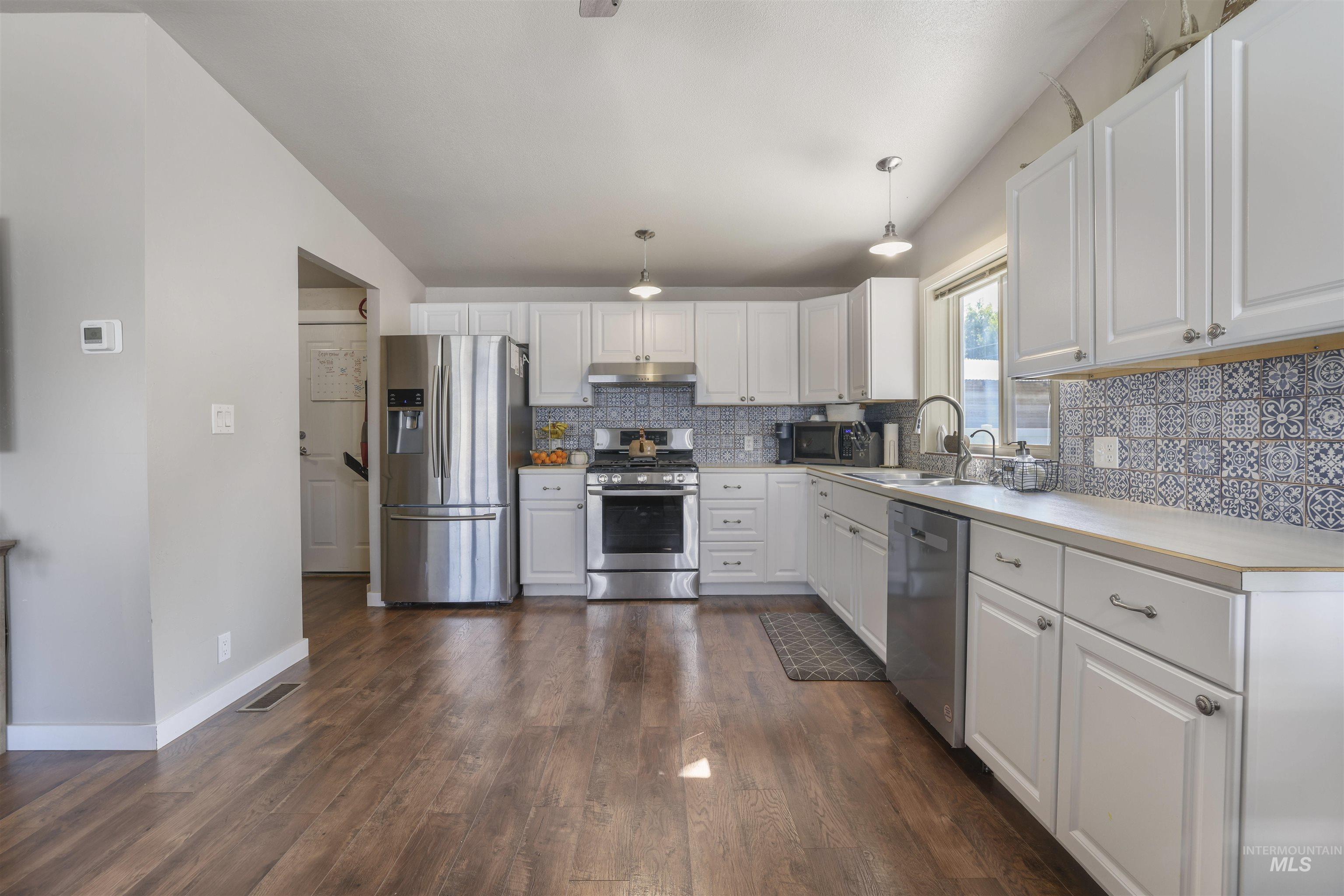 Kitchen featuring light countertops, appliances with stainless steel finishes, white cabinets, decorative light fixtures, and dark wood-style flooring