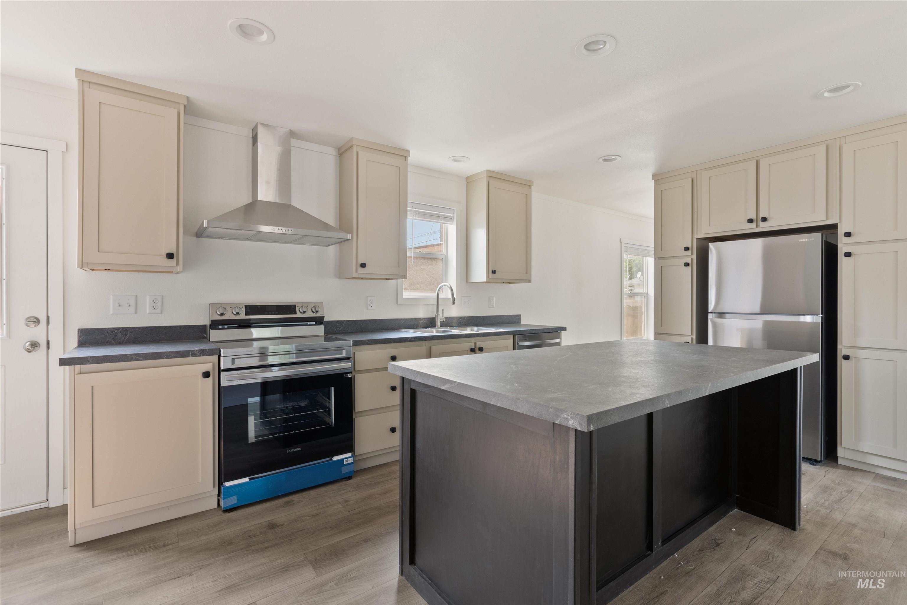 Kitchen featuring cream cabinets, appliances with stainless steel finishes, wall chimney range hood, light wood-style floors, and dark countertops