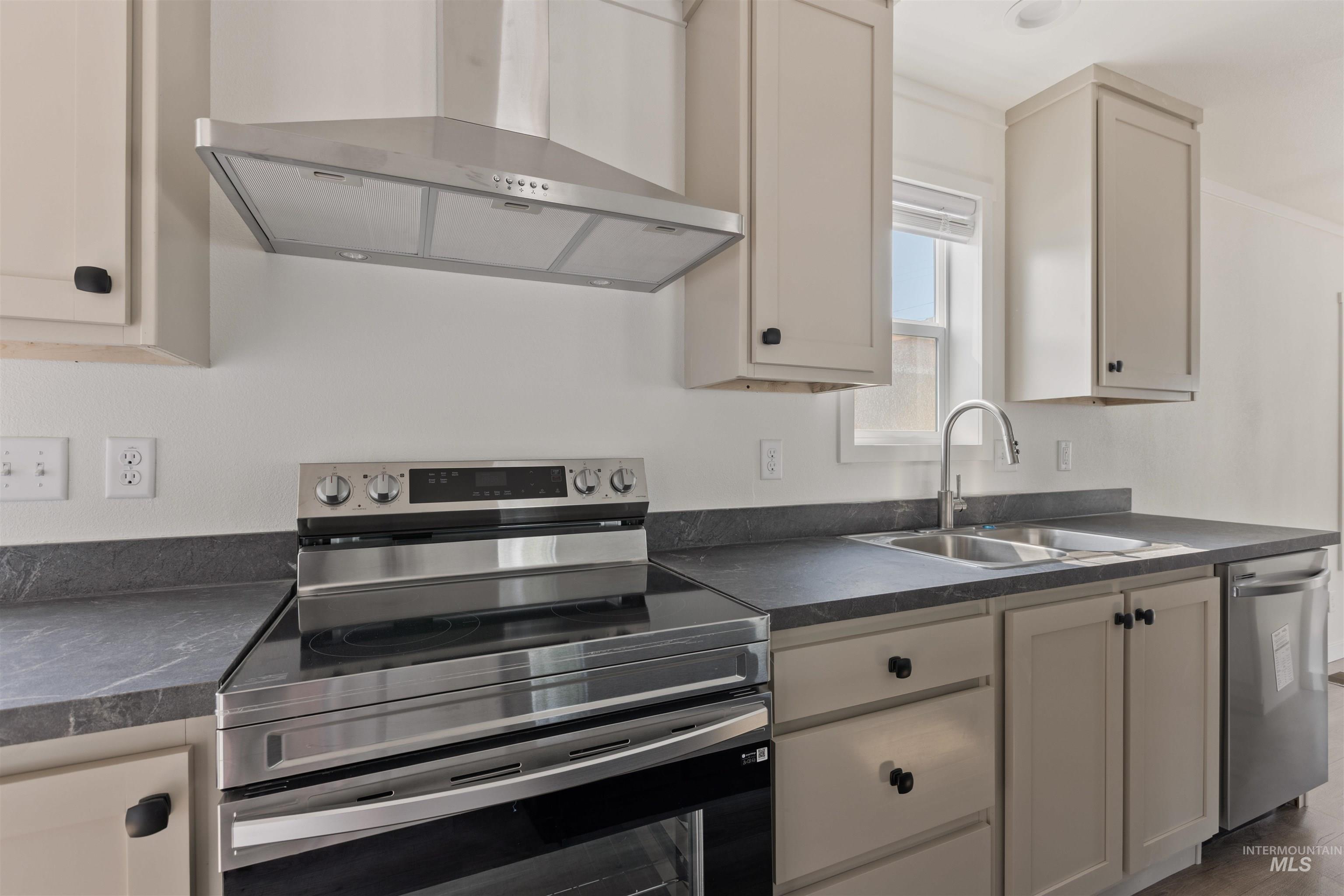 Kitchen with stainless steel appliances, dark countertops, wall chimney range hood, and cream cabinetry