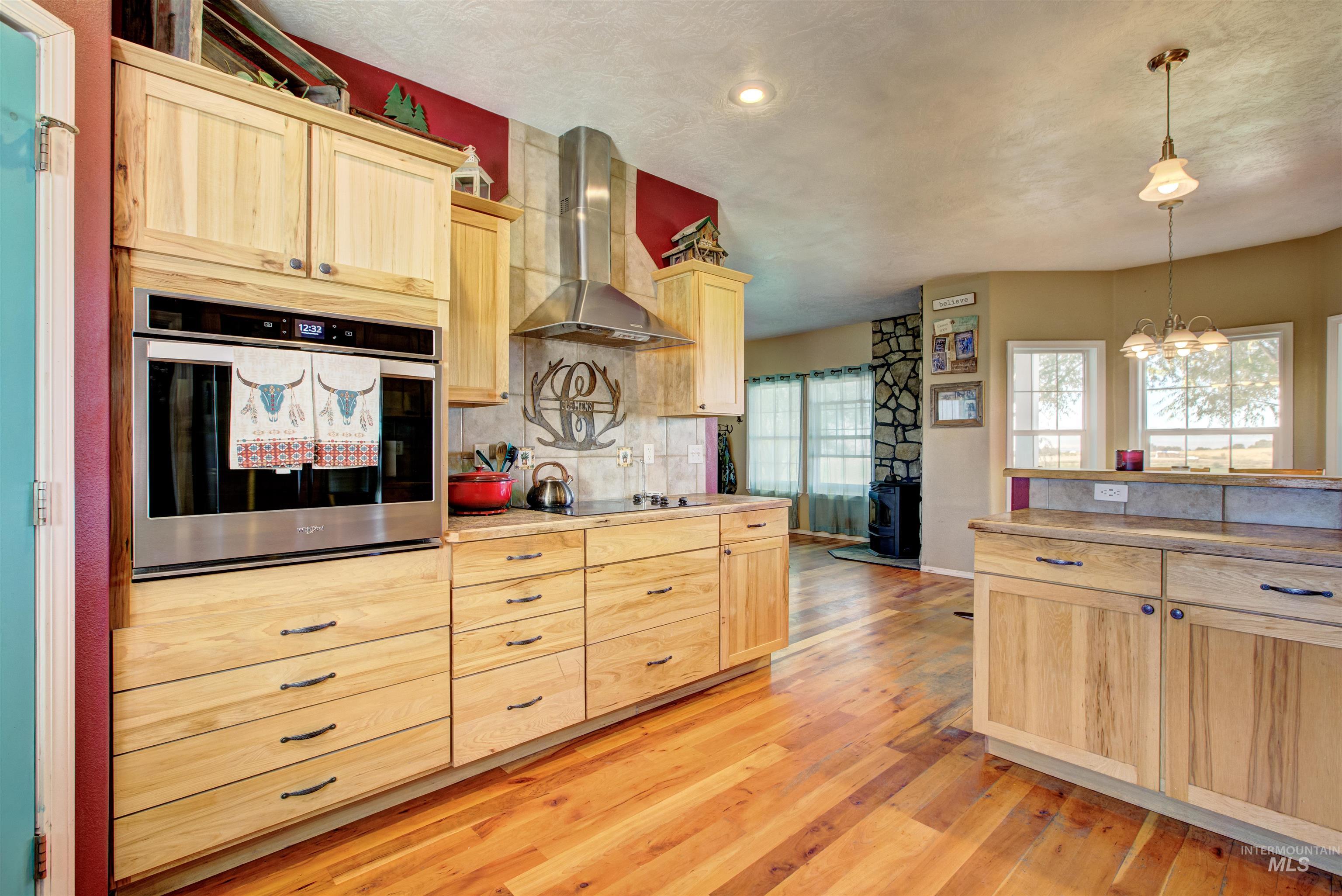 Kitchen featuring light brown cabinetry, stainless steel oven, wall chimney range hood, hanging light fixtures, and backsplash