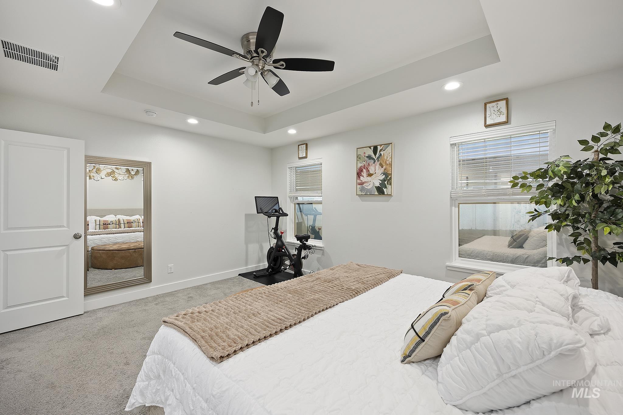 Carpeted bedroom with a tray ceiling, multiple windows, ceiling fan, and recessed lighting