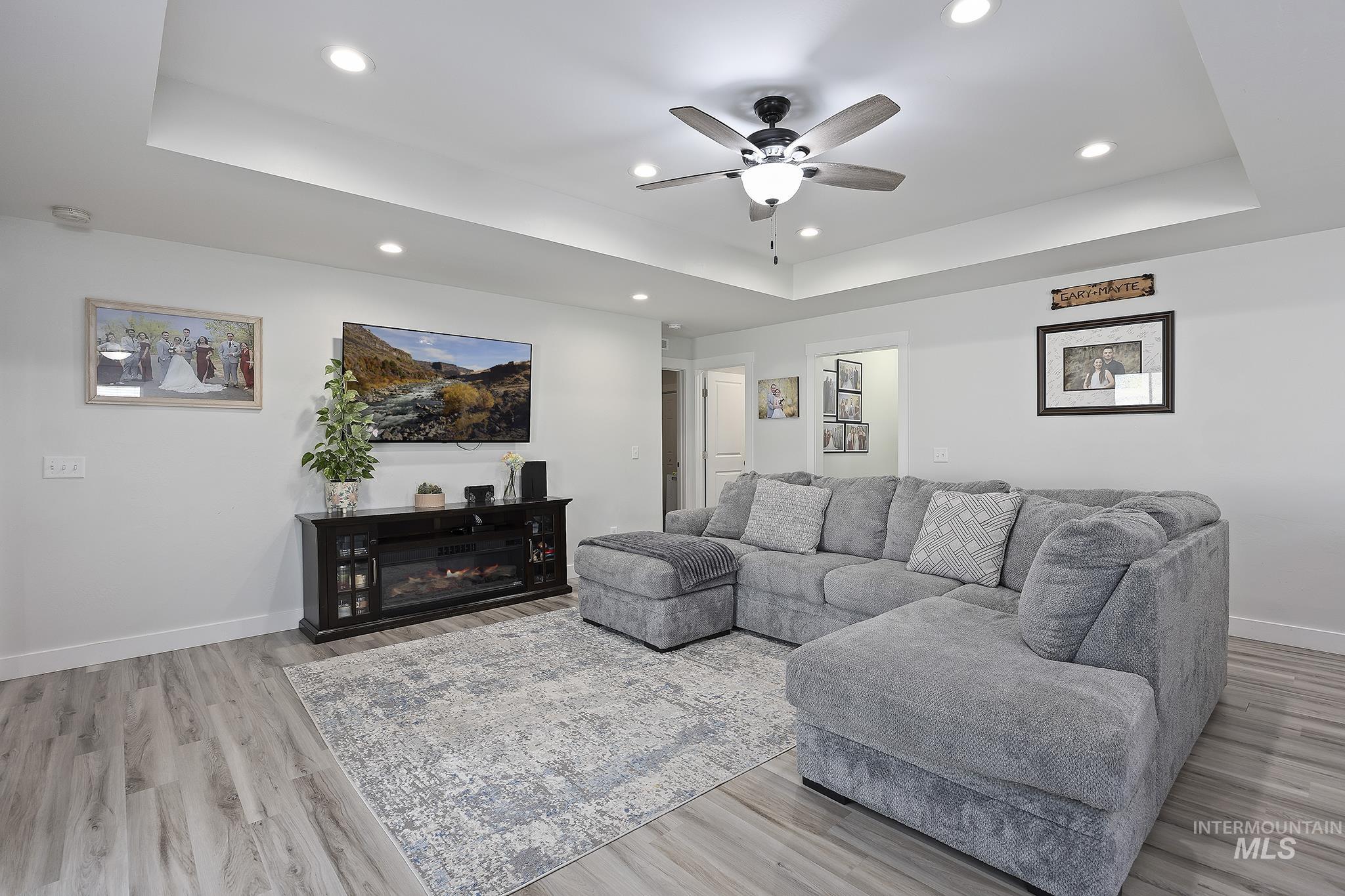 Living area with a raised ceiling, recessed lighting, a glass covered fireplace, light wood finished floors, and ceiling fan