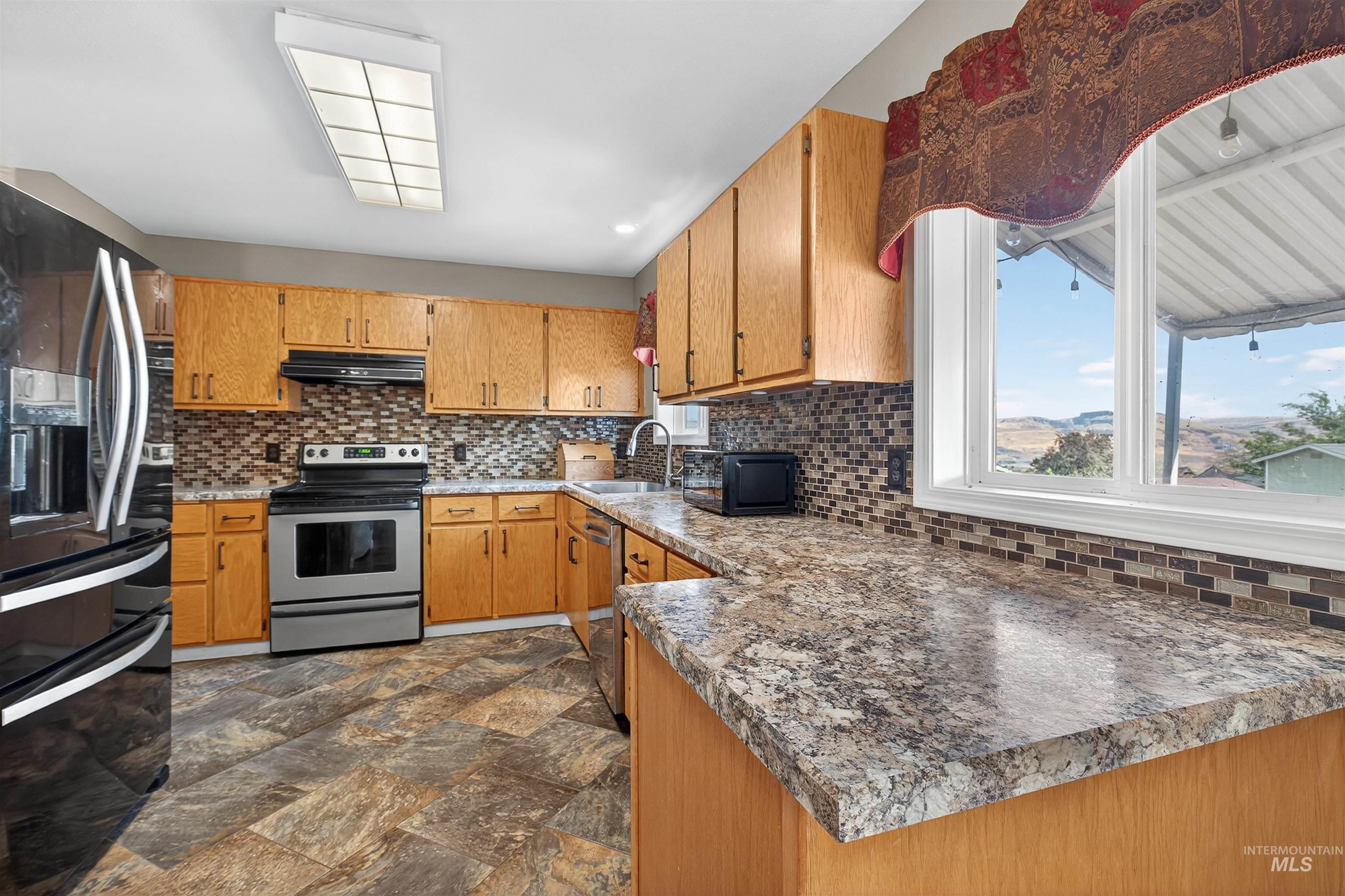 Kitchen with black appliances, decorative backsplash, brown cabinetry, and dark stone finish flooring