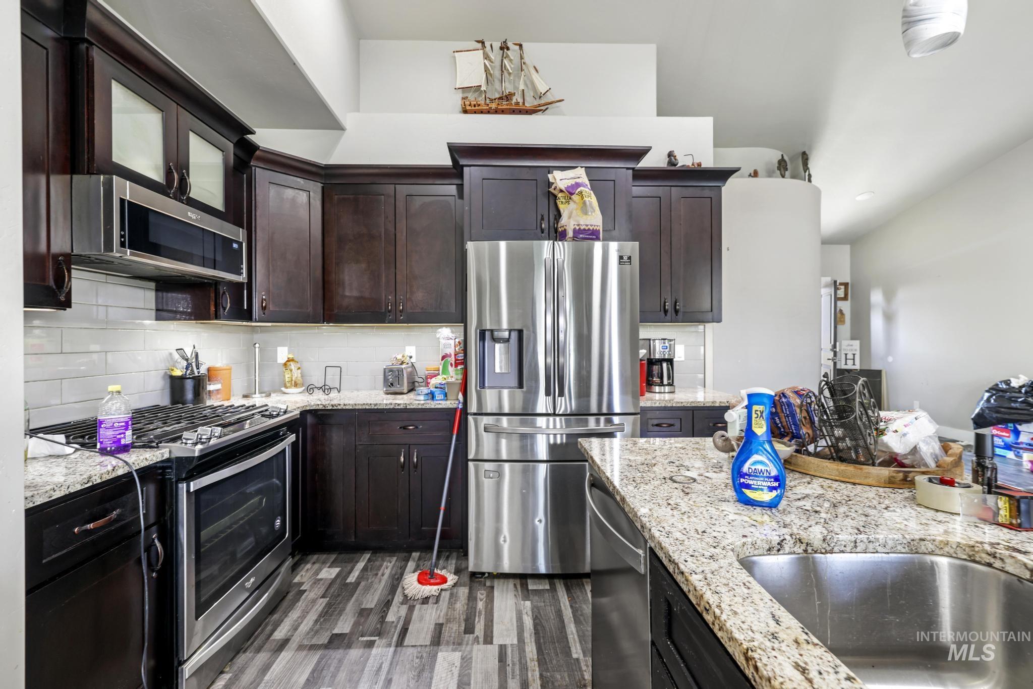 Kitchen featuring stainless steel appliances, dark wood finished floors, tasteful backsplash, light stone counters, and dark brown cabinetry