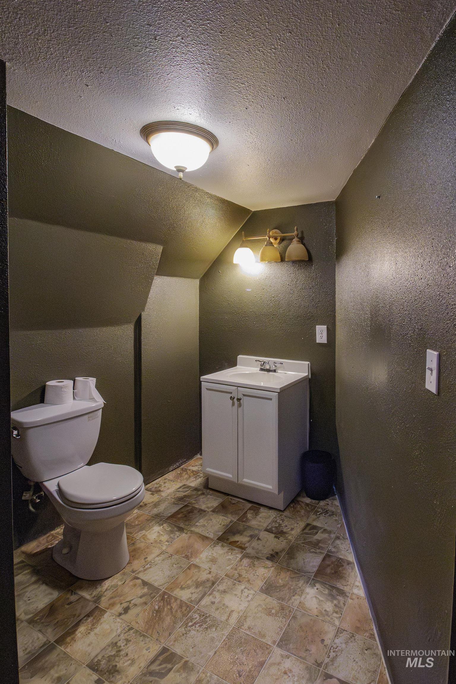 Bathroom with a textured wall, a textured ceiling, vaulted ceiling, stone finish flooring, and vanity