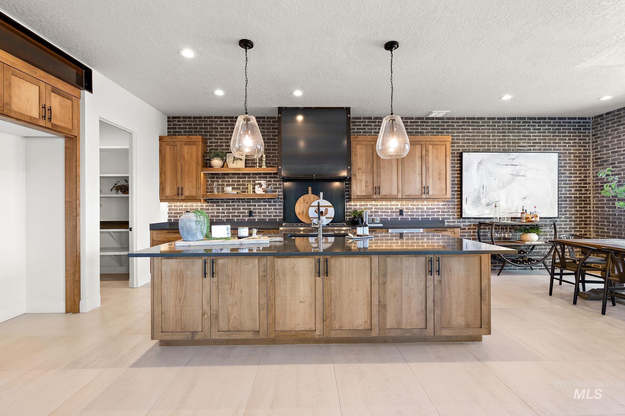 Kitchen featuring open shelves, brown cabinets, a center island with sink, a textured ceiling, and decorative light fixtures