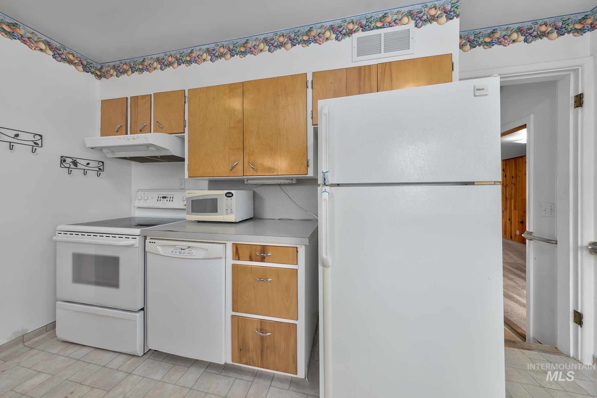 Kitchen with white appliances, brown cabinetry, light countertops, and under cabinet range hood