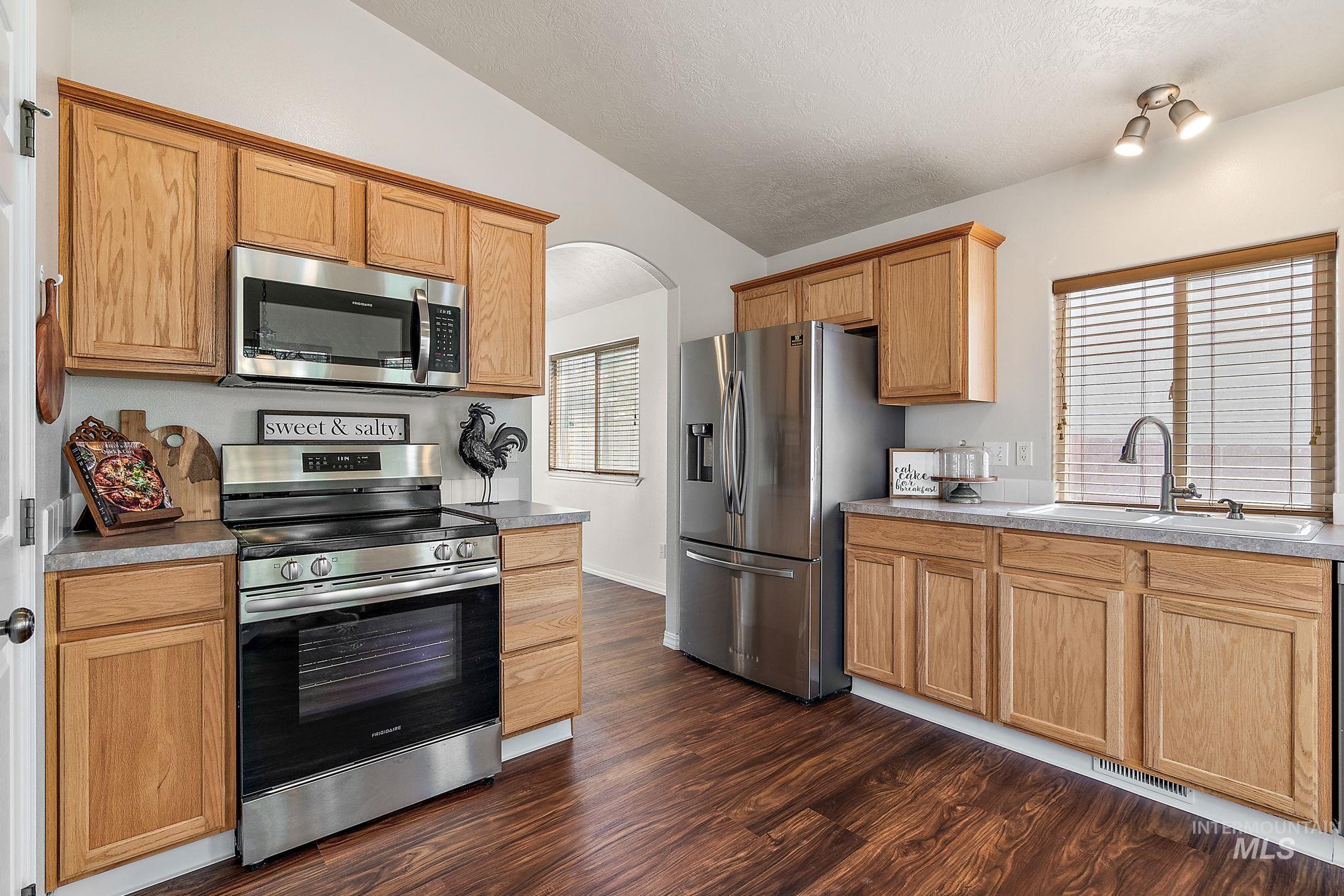 Kitchen with arched walkways, appliances with stainless steel finishes, dark wood-style floors, vaulted ceiling, and light countertops