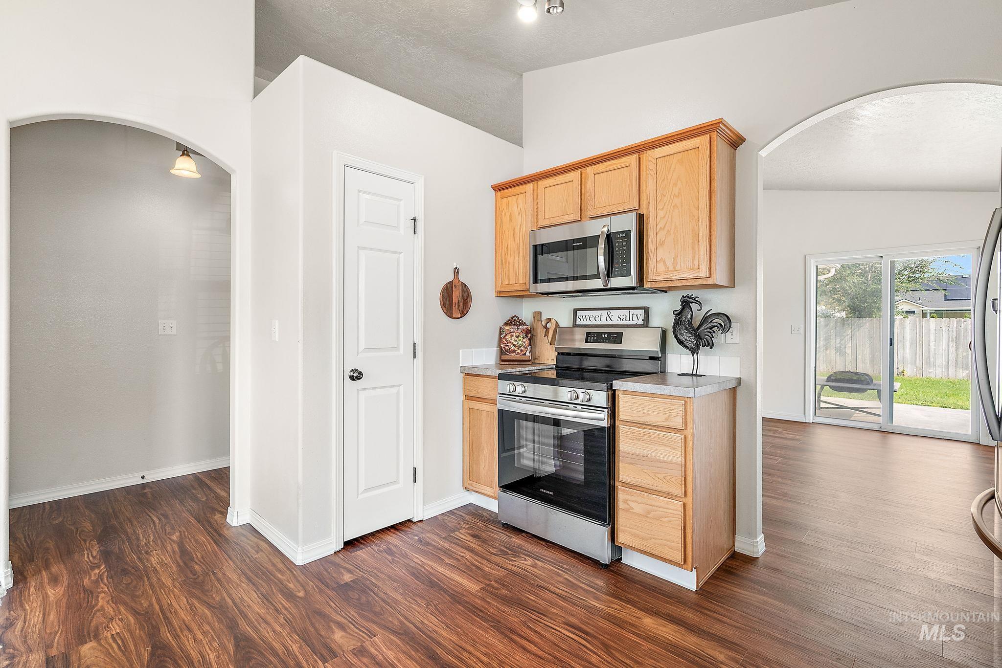 Kitchen with arched walkways, appliances with stainless steel finishes, light countertops, dark wood-style floors, and light brown cabinetry