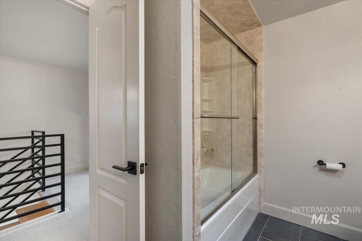 Bathroom featuring shower / bath combination with glass door, a textured wall, and dark tile patterned flooring
