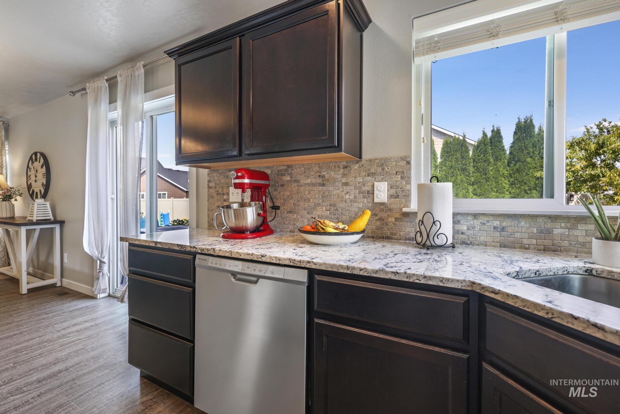 Kitchen featuring dishwasher, backsplash, light stone counters, dark brown cabinets, and dark wood finished floors