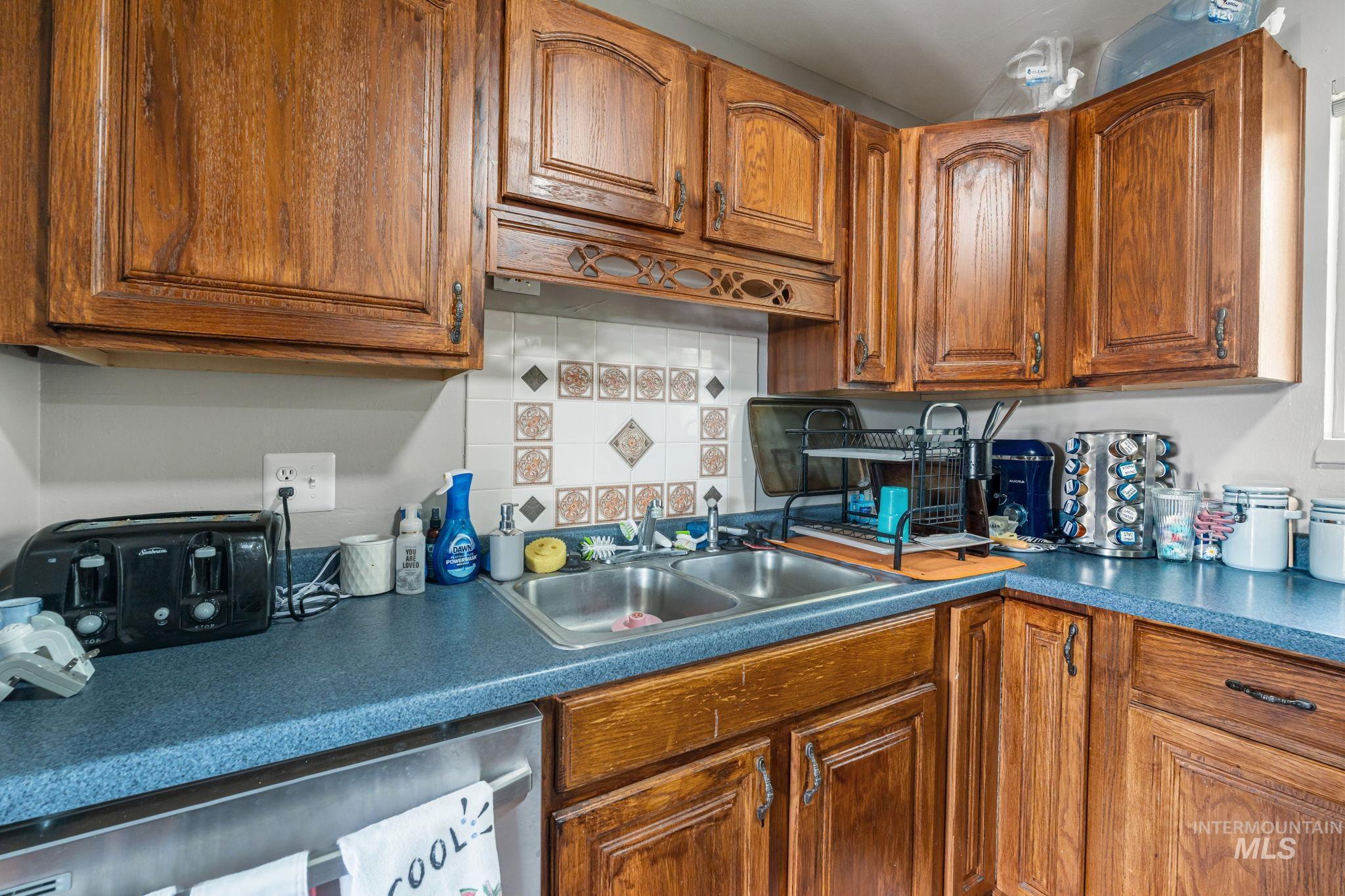 Kitchen with stainless steel dishwasher, brown cabinets, dark countertops, and tasteful backsplash