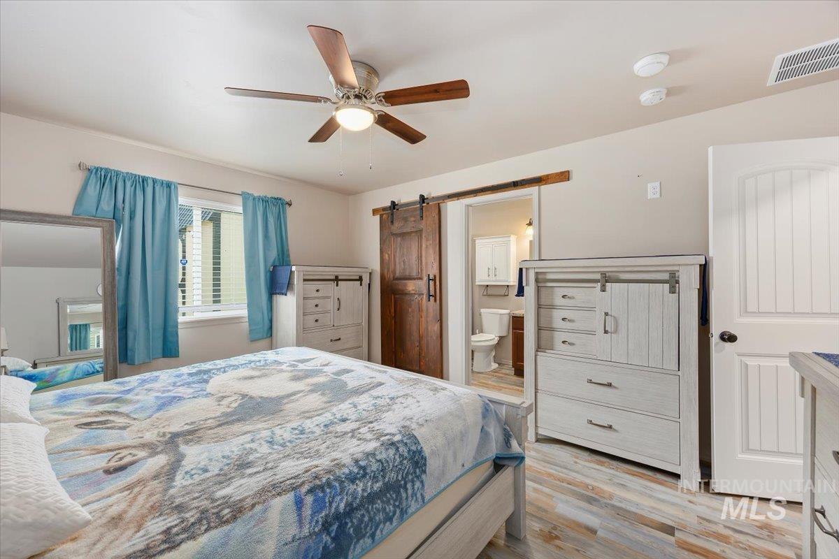 Bedroom with light wood-style flooring, a barn door, ceiling fan, and ensuite bathroom