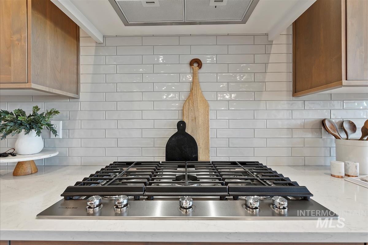 Kitchen view of tasteful backsplash, exhaust hood, stainless steel gas cooktop, and light stone counters
