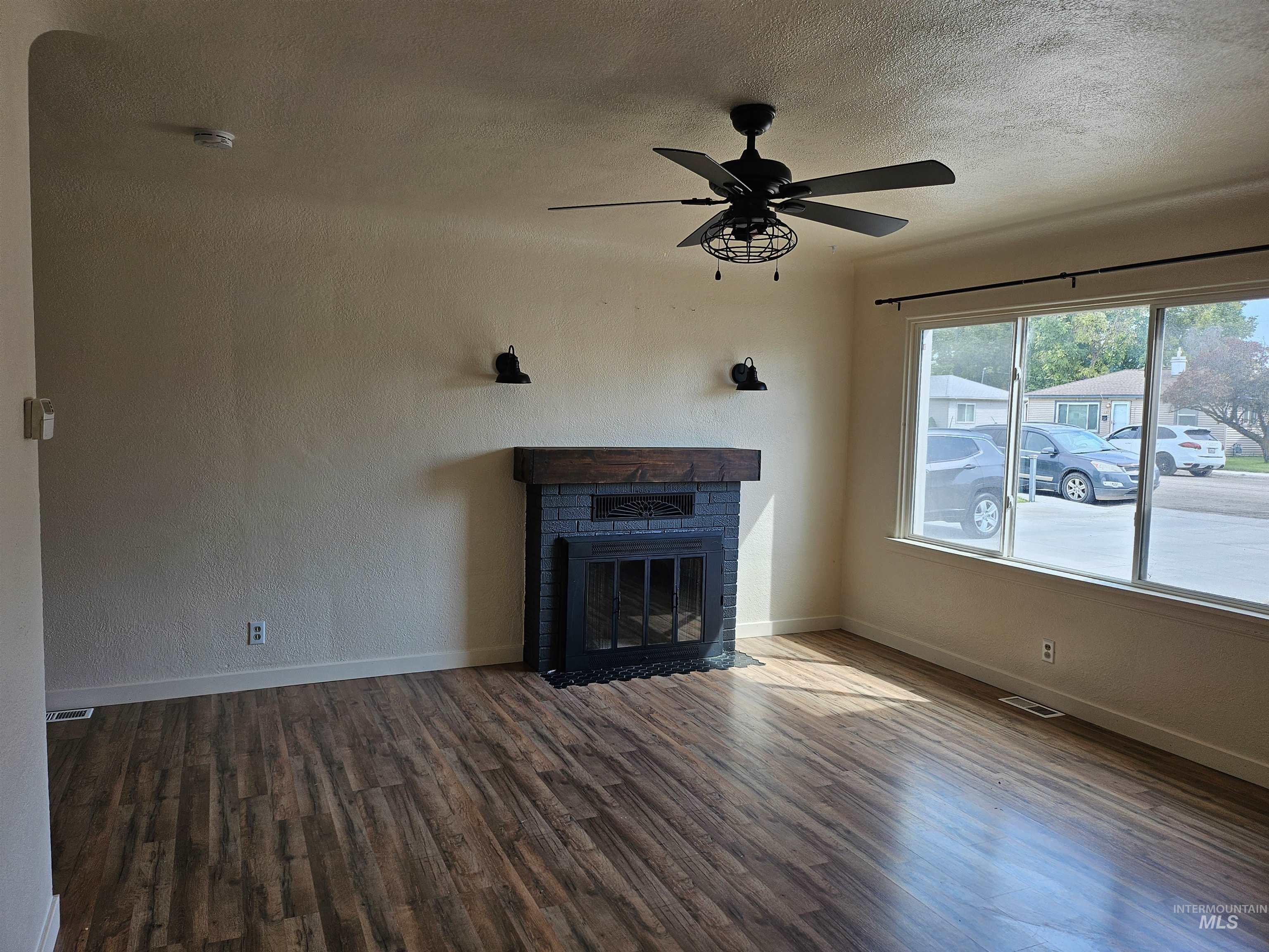 Unfurnished living room with a brick fireplace, a textured ceiling, dark wood-type flooring, ceiling fan, and a textured wall