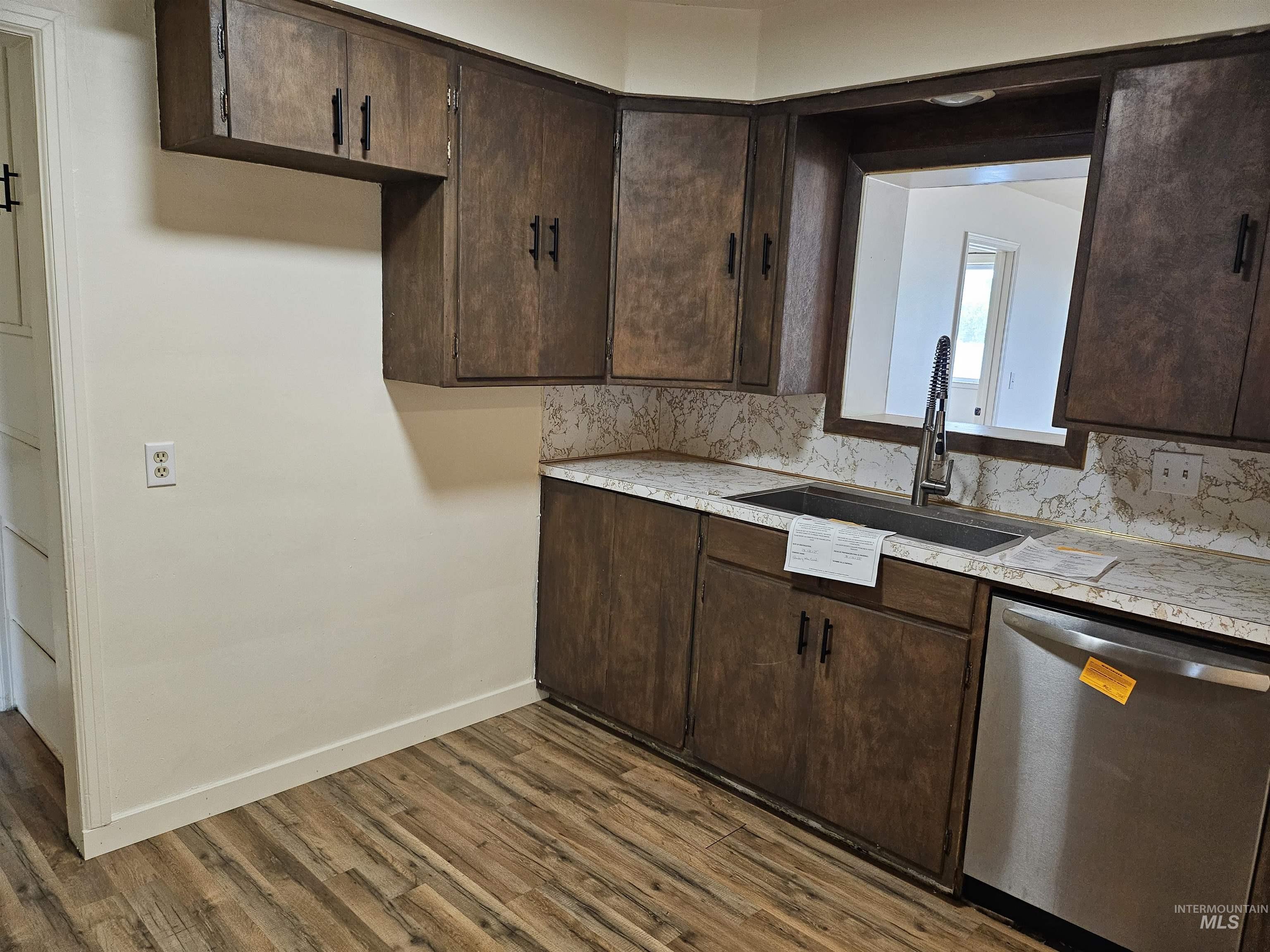 Kitchen with dark brown cabinets, stainless steel dishwasher, light countertops, dark wood-style floors, and tasteful backsplash