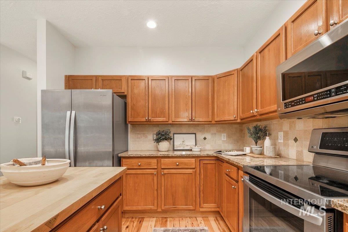 Kitchen with appliances with stainless steel finishes, brown cabinetry, tasteful backsplash, light wood-type flooring, and light stone countertops