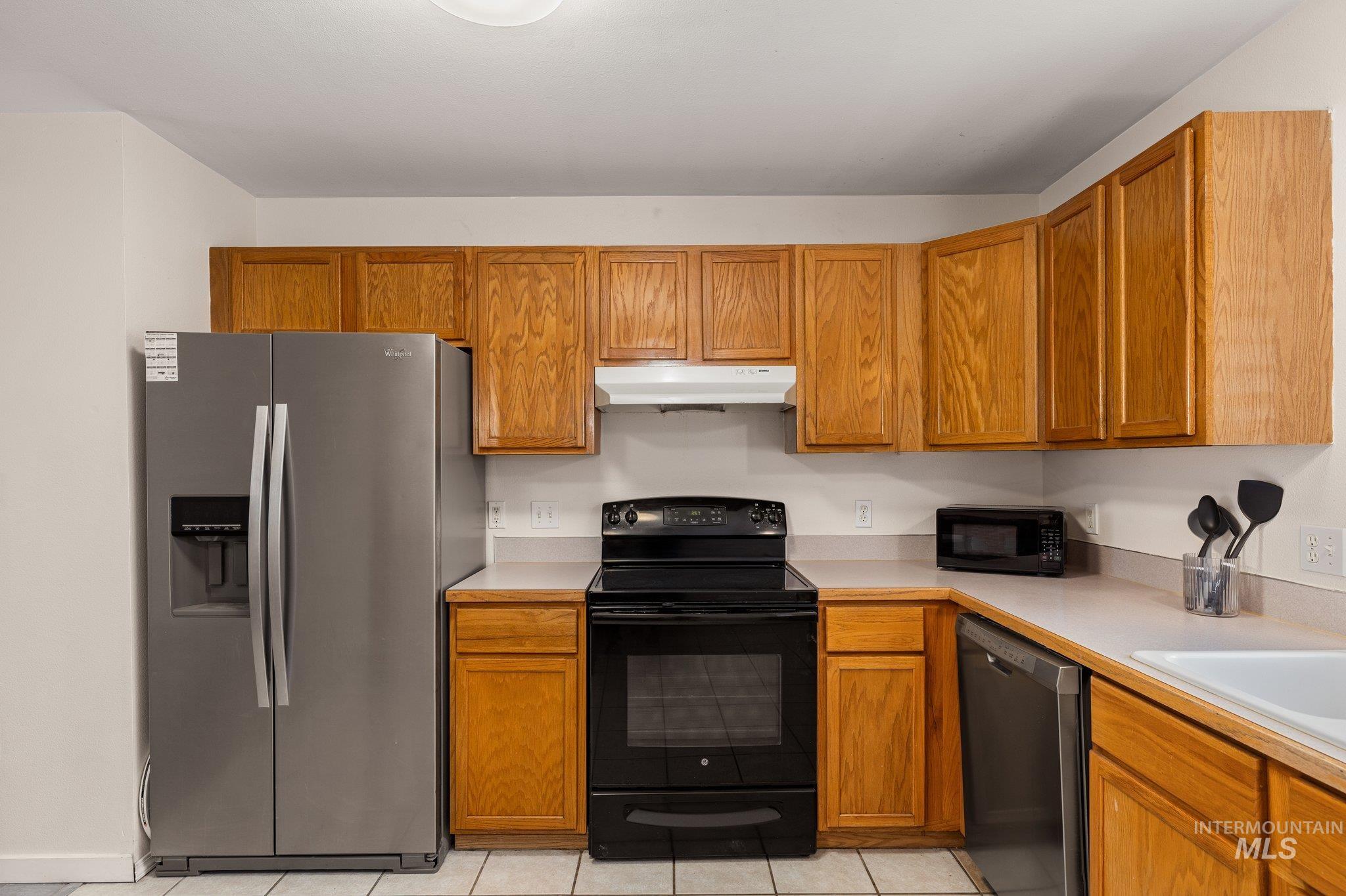 Kitchen featuring black appliances, light countertops, brown cabinets, under cabinet range hood, and light tile patterned flooring
