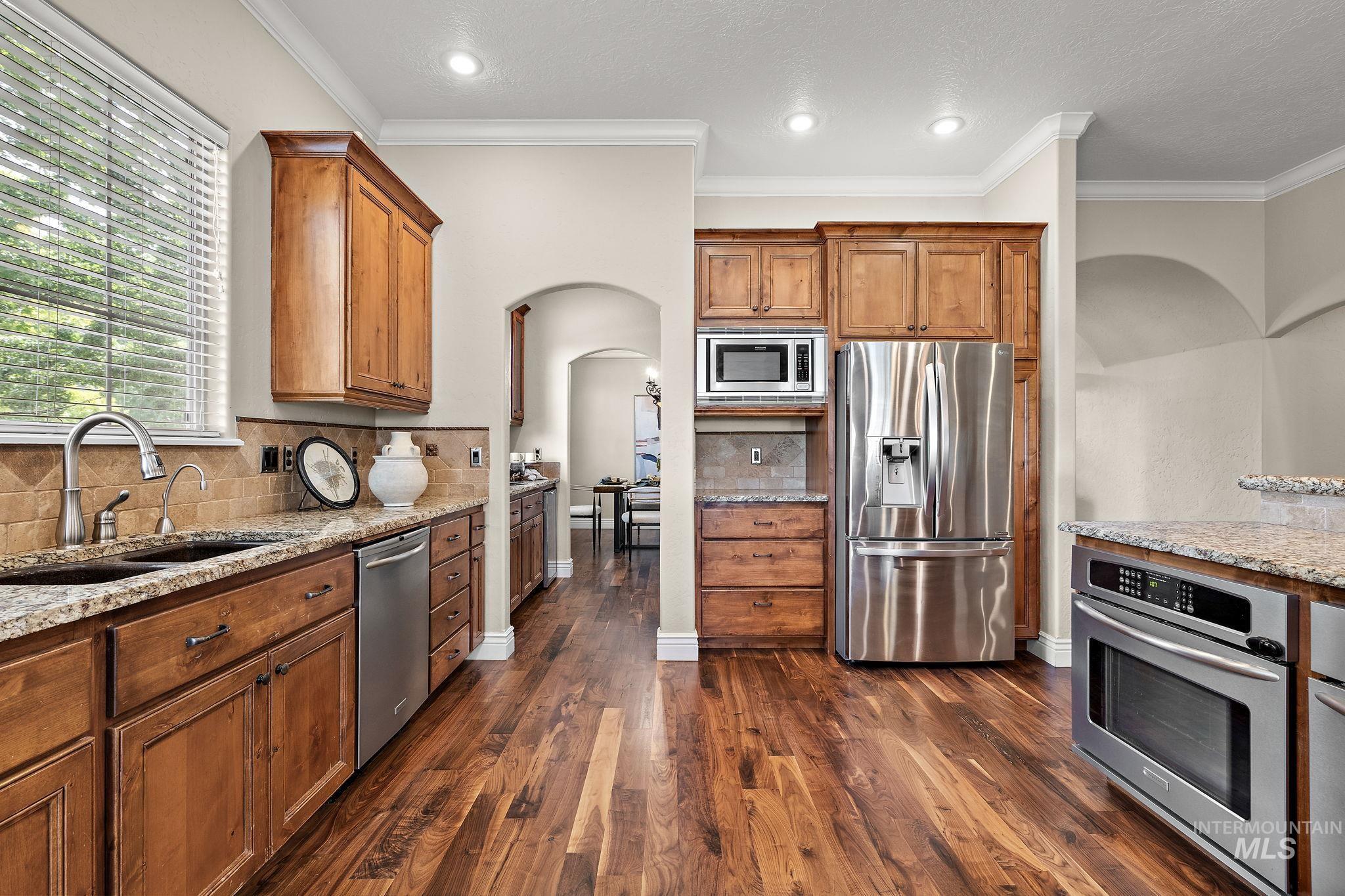 Kitchen featuring arched walkways, stainless steel appliances, light stone countertops, brown cabinetry, and dark wood-style flooring