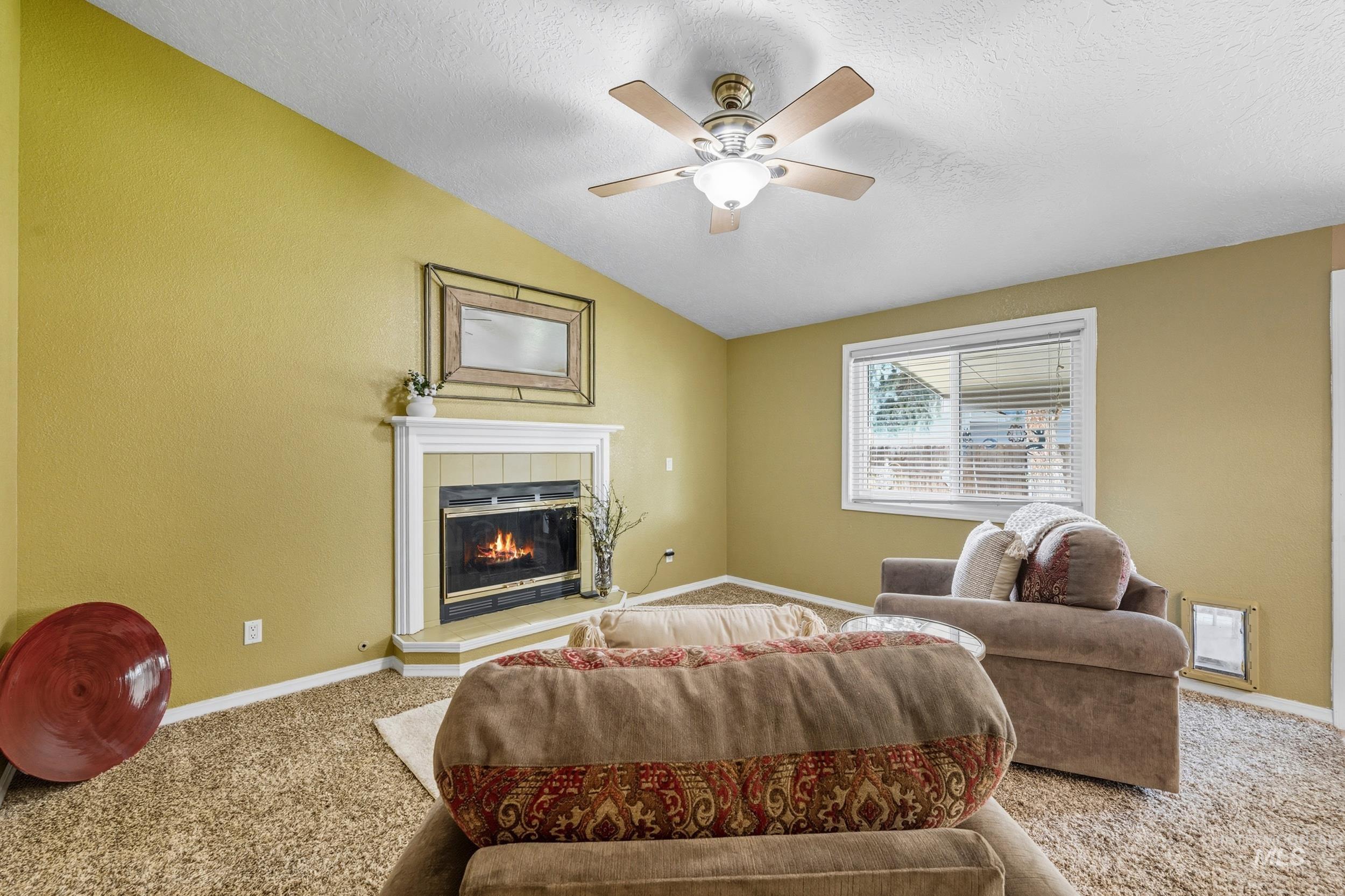 Living room with carpet, a tiled fireplace, vaulted ceiling, ceiling fan, and a textured ceiling