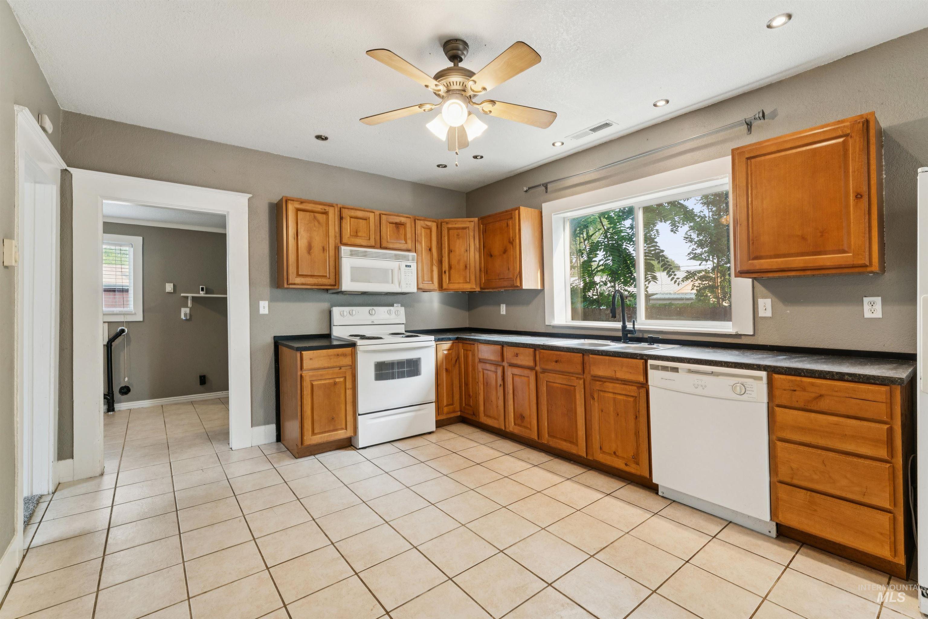 Kitchen featuring dark countertops, white appliances, light tile patterned floors, brown cabinetry, and recessed lighting