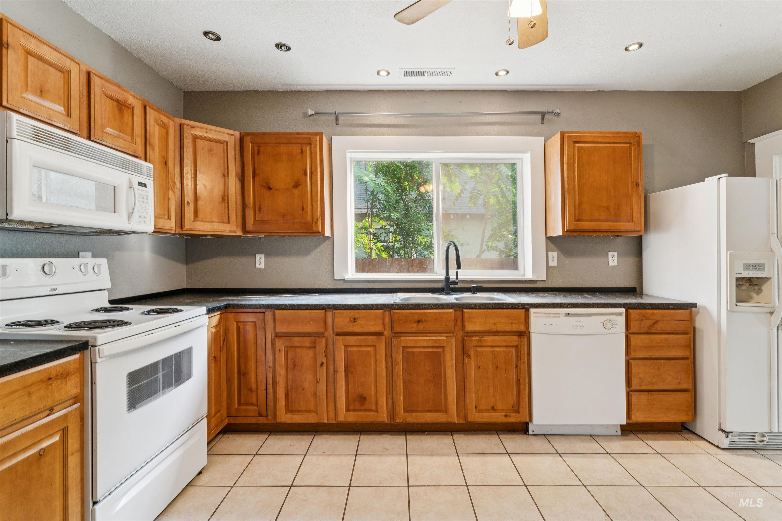 Kitchen with white appliances, light tile patterned floors, dark countertops, brown cabinetry, and recessed lighting