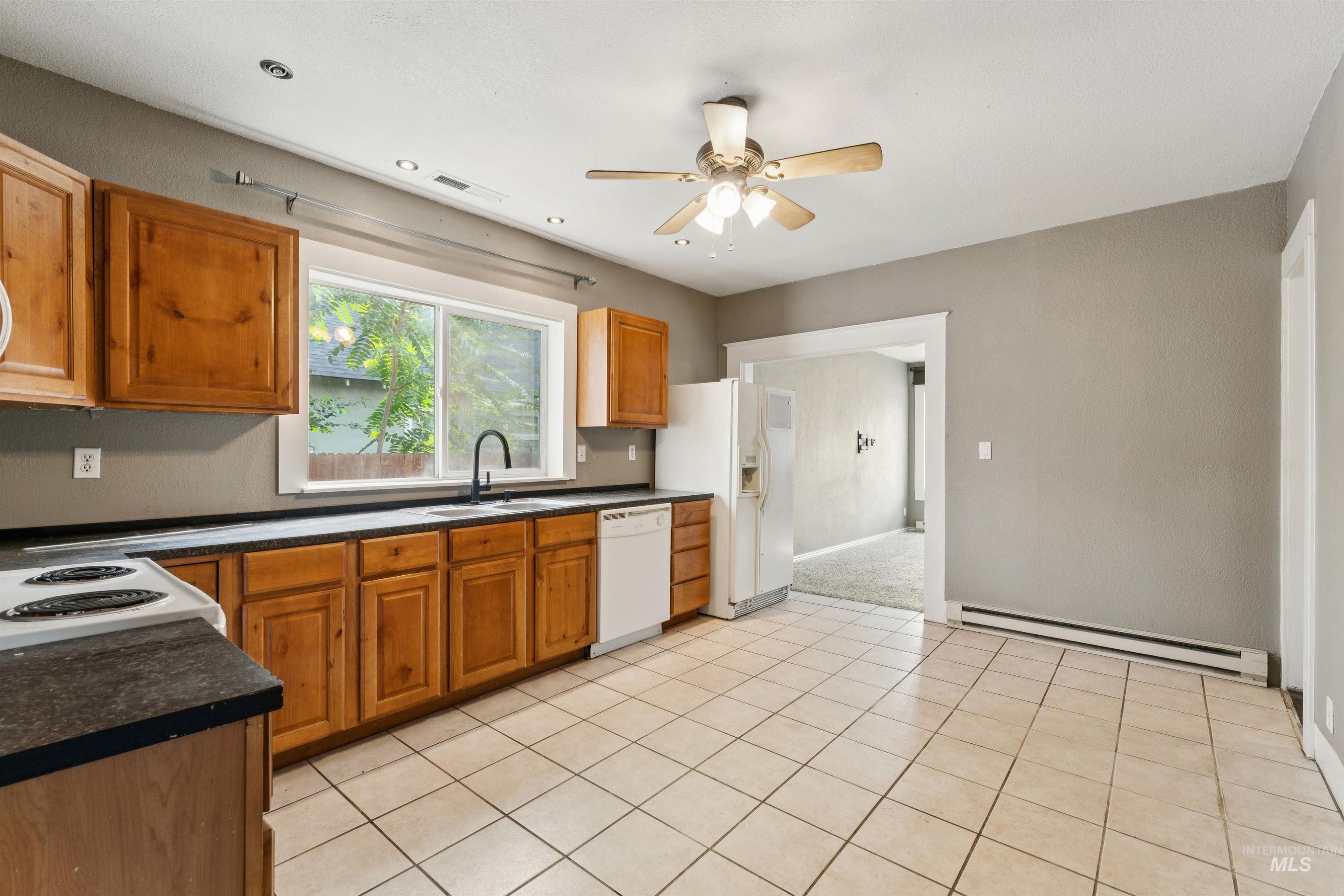 Kitchen with dark countertops, brown cabinetry, baseboard heating, light tile patterned floors, and white appliances