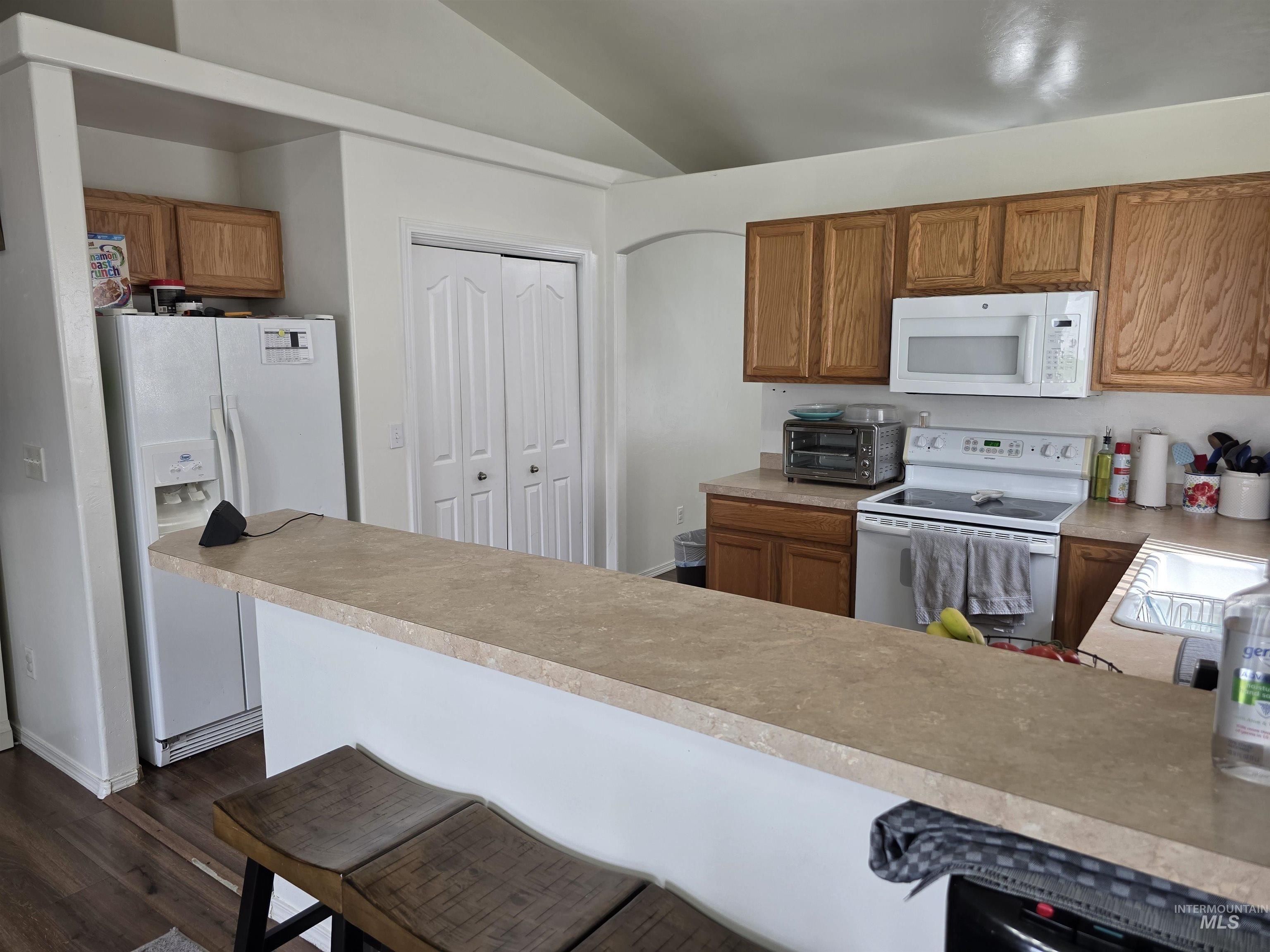 Kitchen featuring brown cabinetry, white appliances, a kitchen breakfast bar, light countertops, and dark wood-type flooring