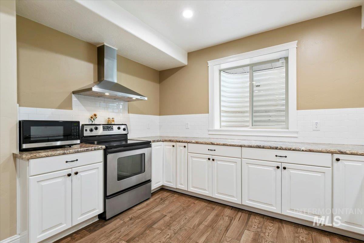 Kitchen featuring appliances with stainless steel finishes, white cabinets, wall chimney range hood, light wood finished floors, and light stone countertops