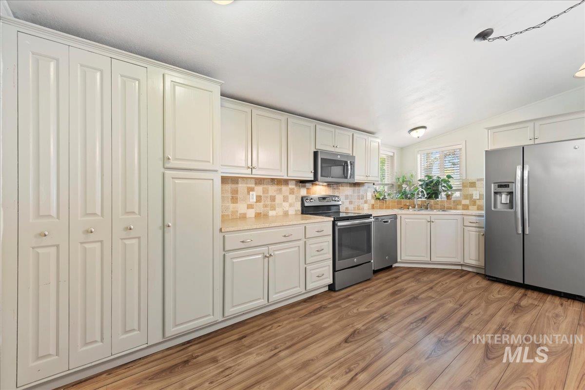 Kitchen with tasteful backsplash, appliances with stainless steel finishes, vaulted ceiling, light wood-style floors, and white cabinetry