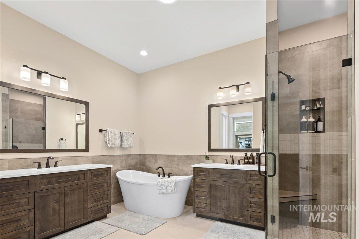 Full bathroom featuring tile walls, a stall shower, two vanities, a freestanding tub, and wainscoting