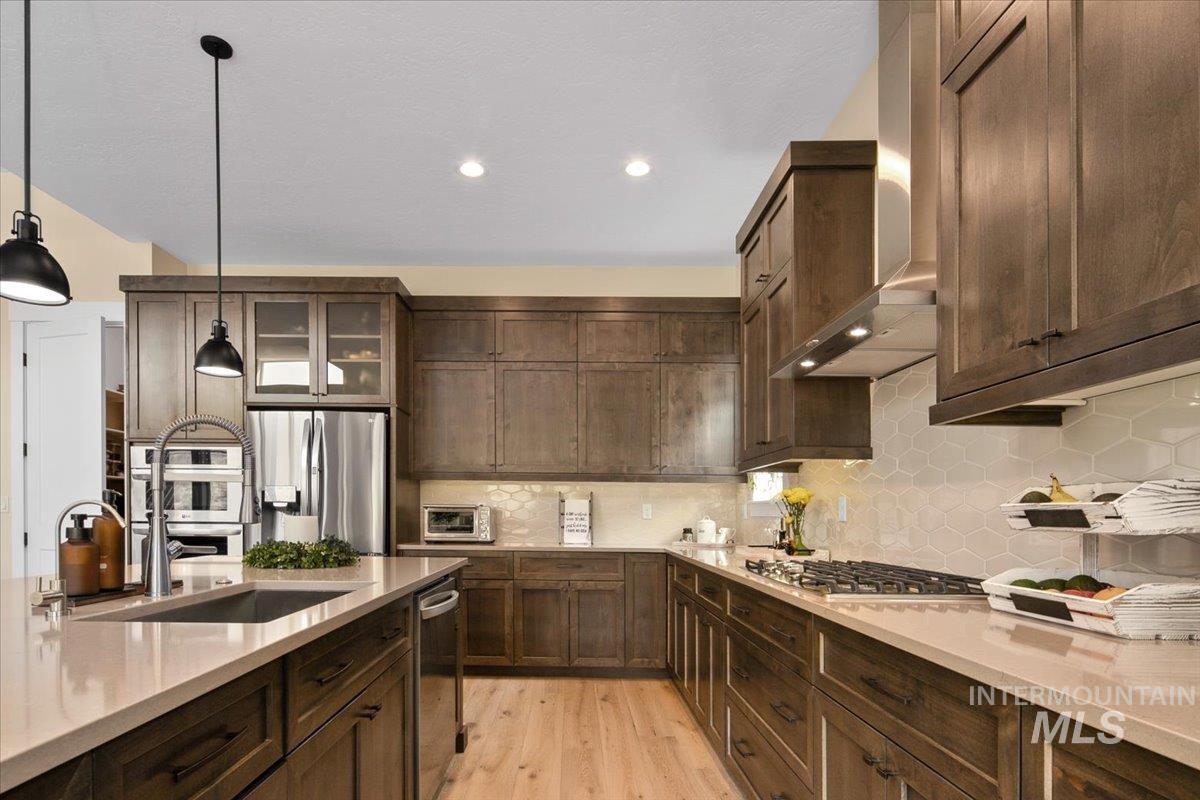 Kitchen featuring dark brown cabinets, backsplash, light wood-type flooring, appliances with stainless steel finishes, and hanging light fixtures