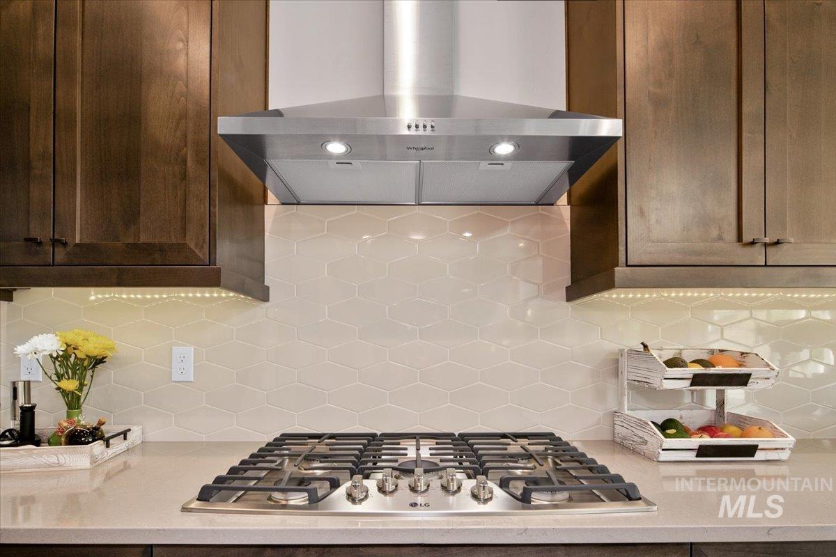 Kitchen featuring wall chimney range hood, backsplash, stainless steel gas cooktop, light stone counters, and dark brown cabinets