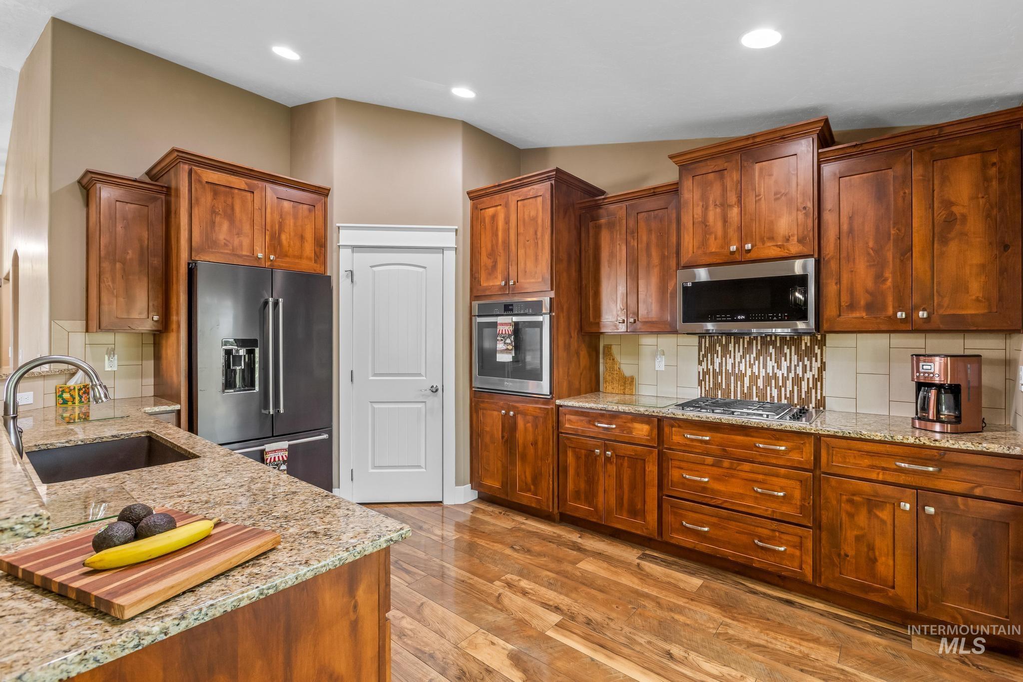 Kitchen with decorative backsplash, appliances with stainless steel finishes, light stone countertops, light wood-style flooring, and brown cabinetry