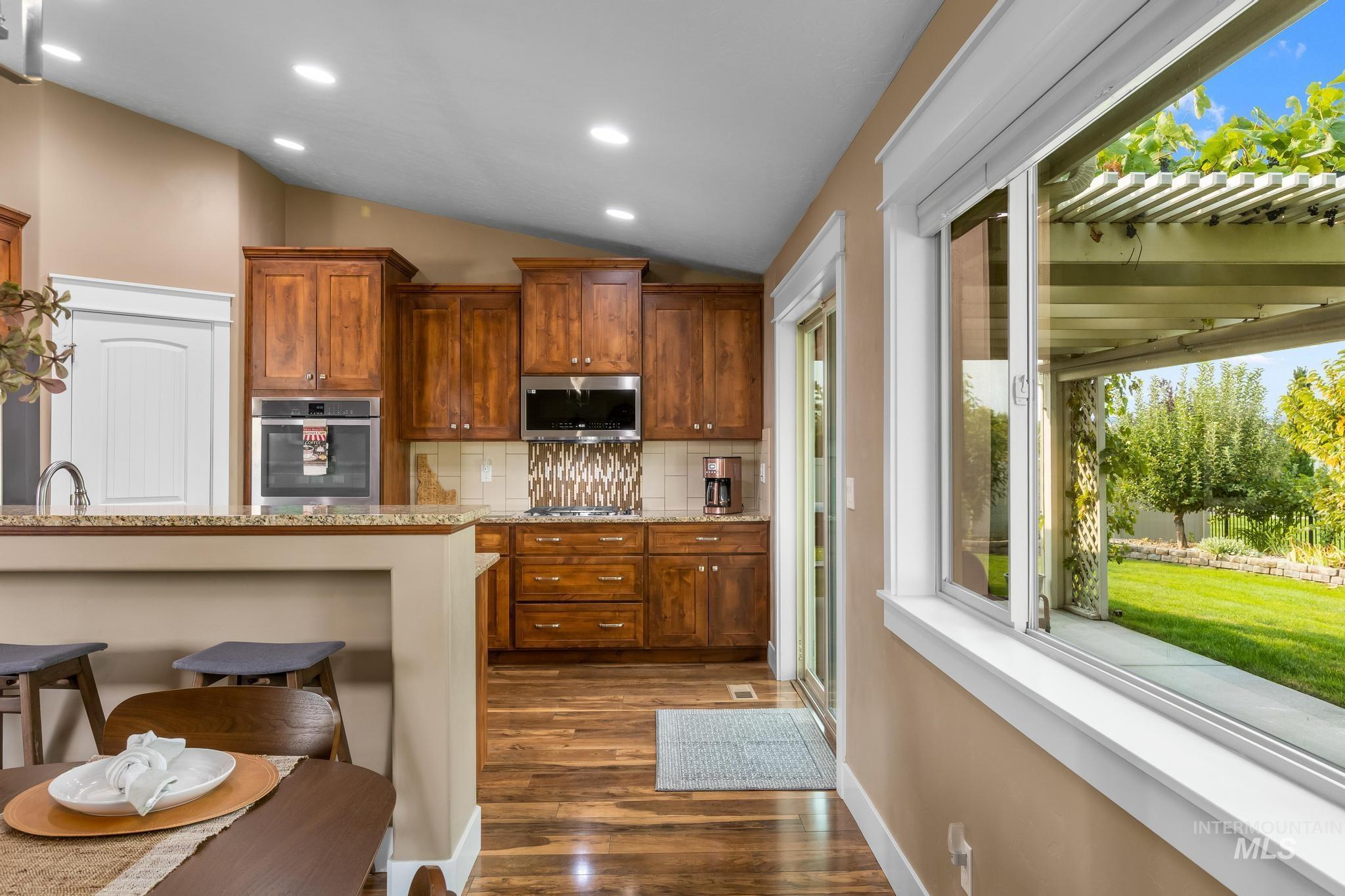 Kitchen featuring brown cabinetry, vaulted ceiling, light stone countertops, backsplash, and a kitchen breakfast bar