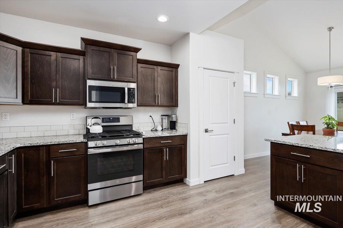 Kitchen with appliances with stainless steel finishes, dark brown cabinets, light stone counters, light wood-style flooring, and lofted ceiling