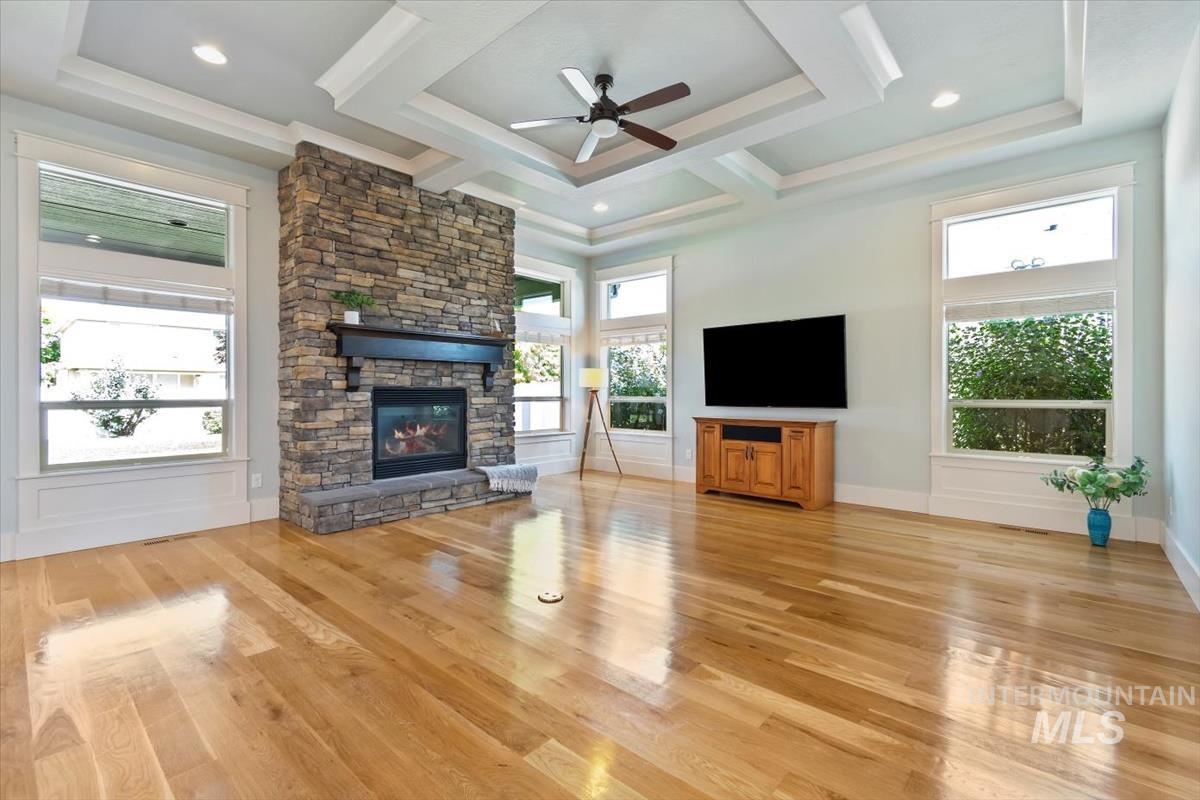 Unfurnished living room with a ceiling fan, light wood-style floors, a stone fireplace, coffered ceiling, and beam ceiling