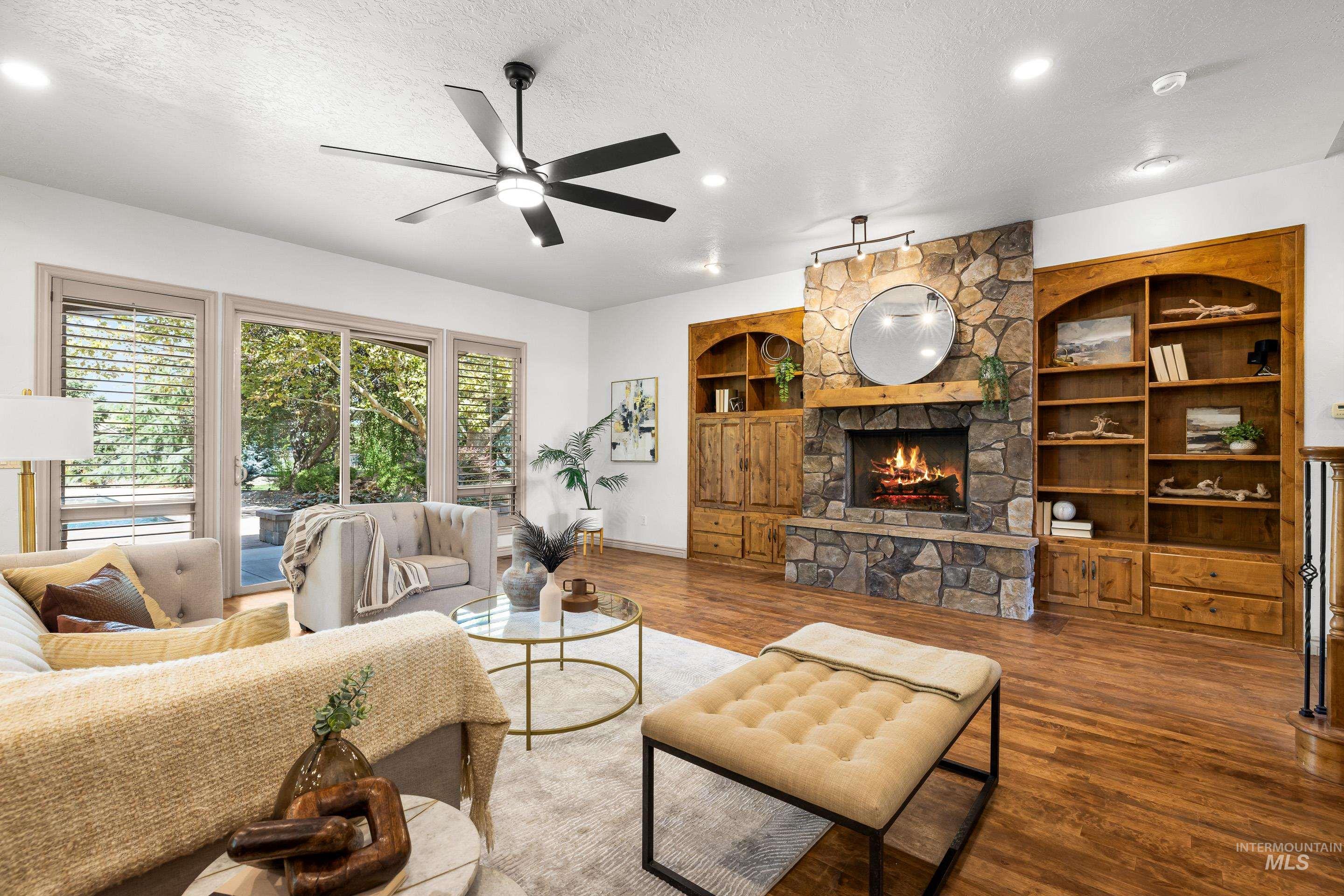 Living room with a textured ceiling, wood finished floors, built in features, a ceiling fan, and a stone fireplace