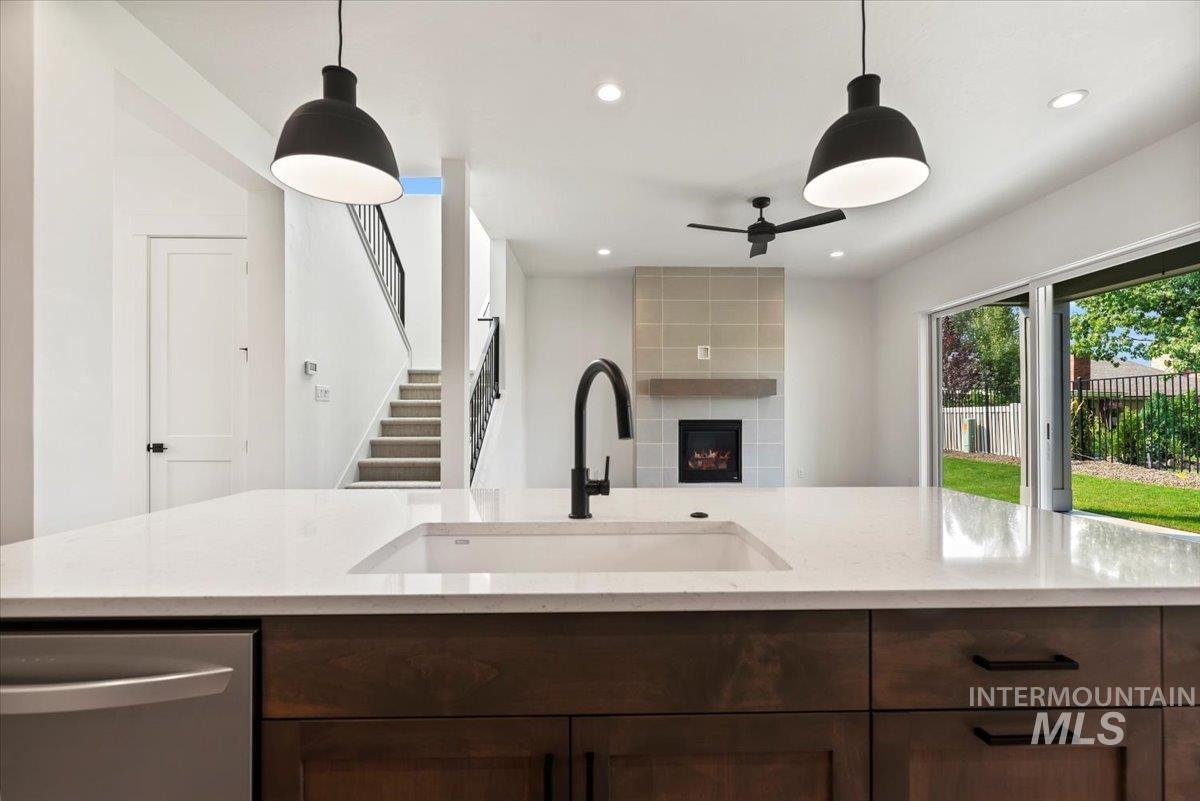 Kitchen featuring dark brown cabinets, stainless steel dishwasher, a fireplace, hanging light fixtures, and light stone counters