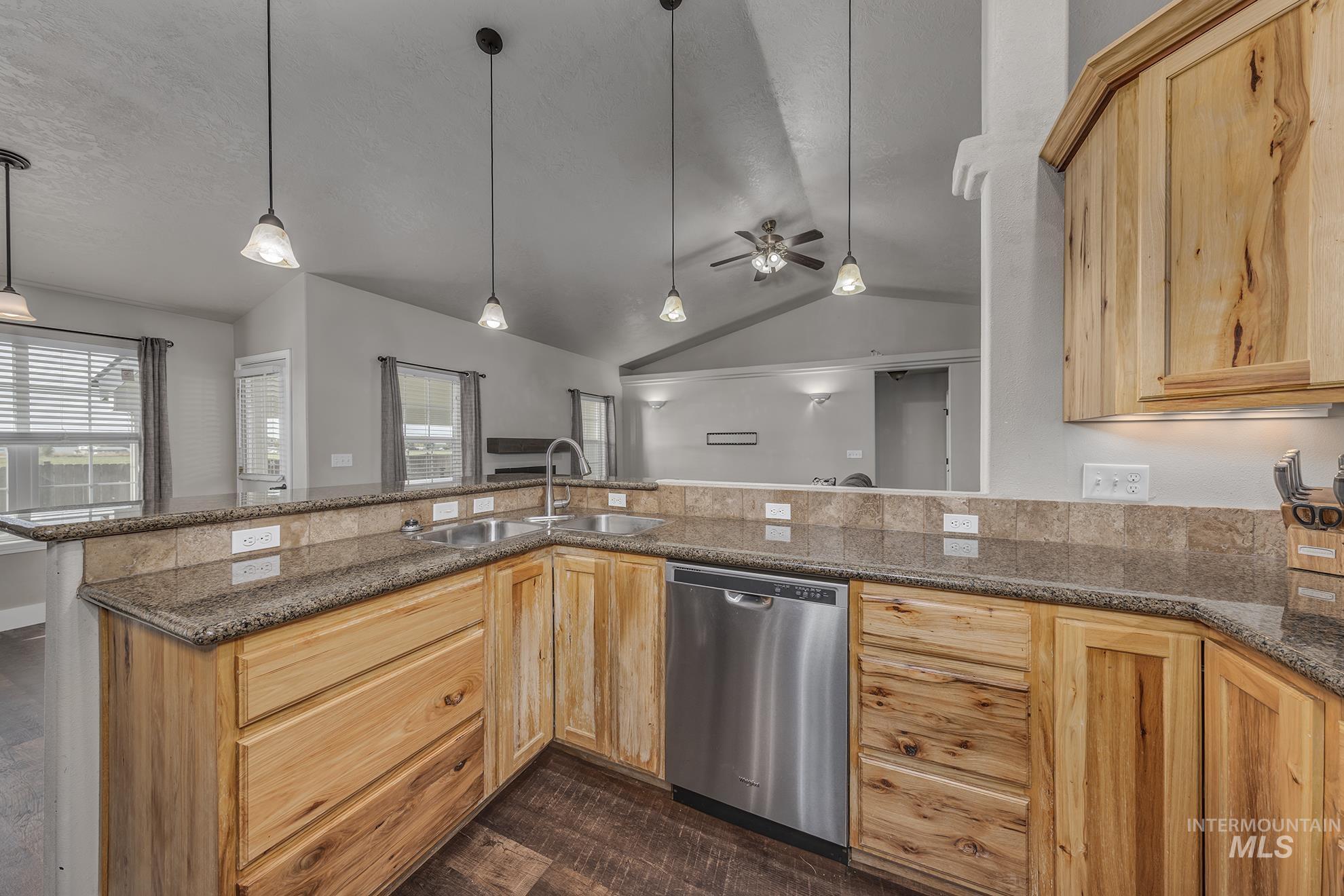 Kitchen with vaulted ceiling, light brown cabinets, pendant lighting, stainless steel dishwasher, and dark stone countertops