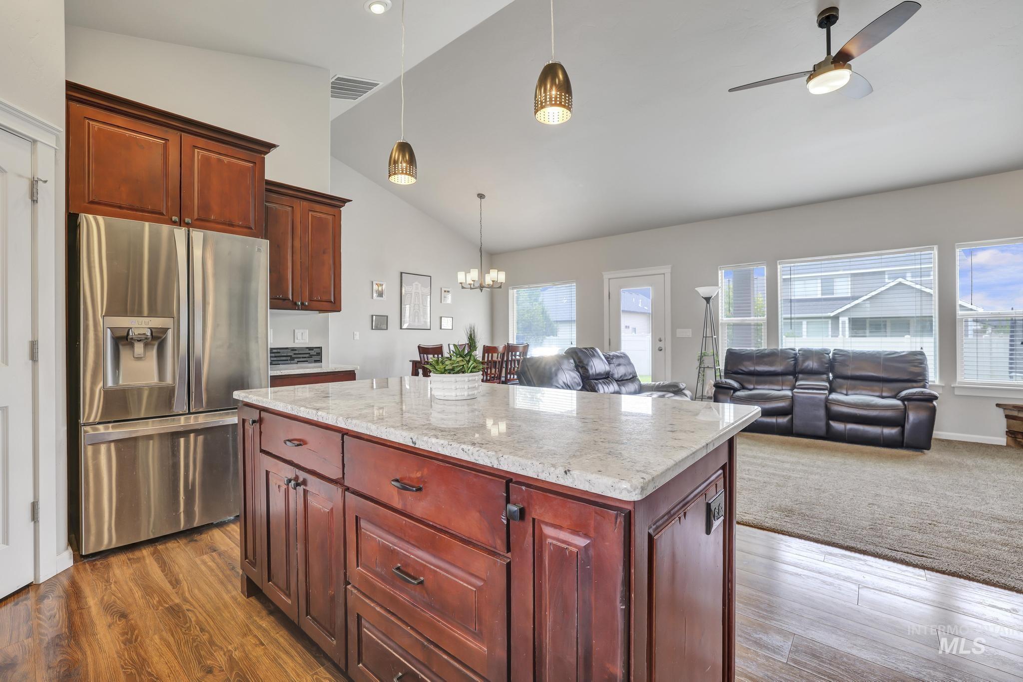 Kitchen with stainless steel fridge, lofted ceiling, light stone counters, pendant lighting, and dark brown cabinets