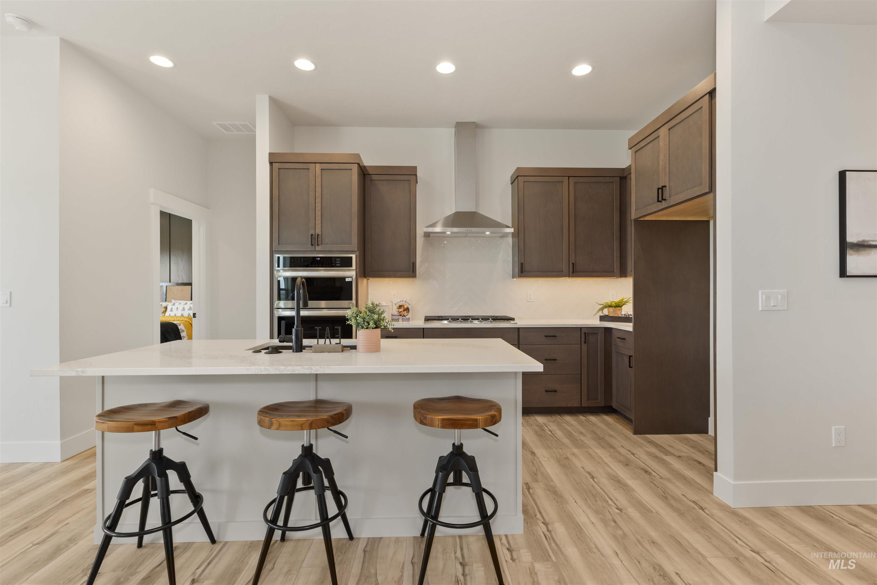 Kitchen featuring recessed lighting, a center island with sink, light wood-style floors, a breakfast bar area, and wall chimney exhaust hood