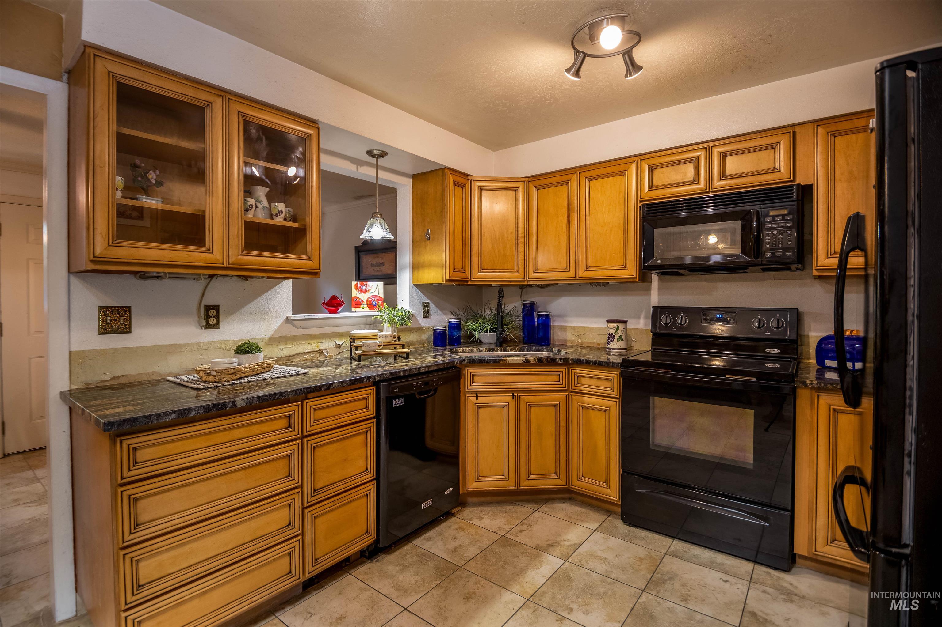 Kitchen featuring brown cabinetry, black appliances, pendant lighting, light tile patterned flooring, and glass insert cabinets